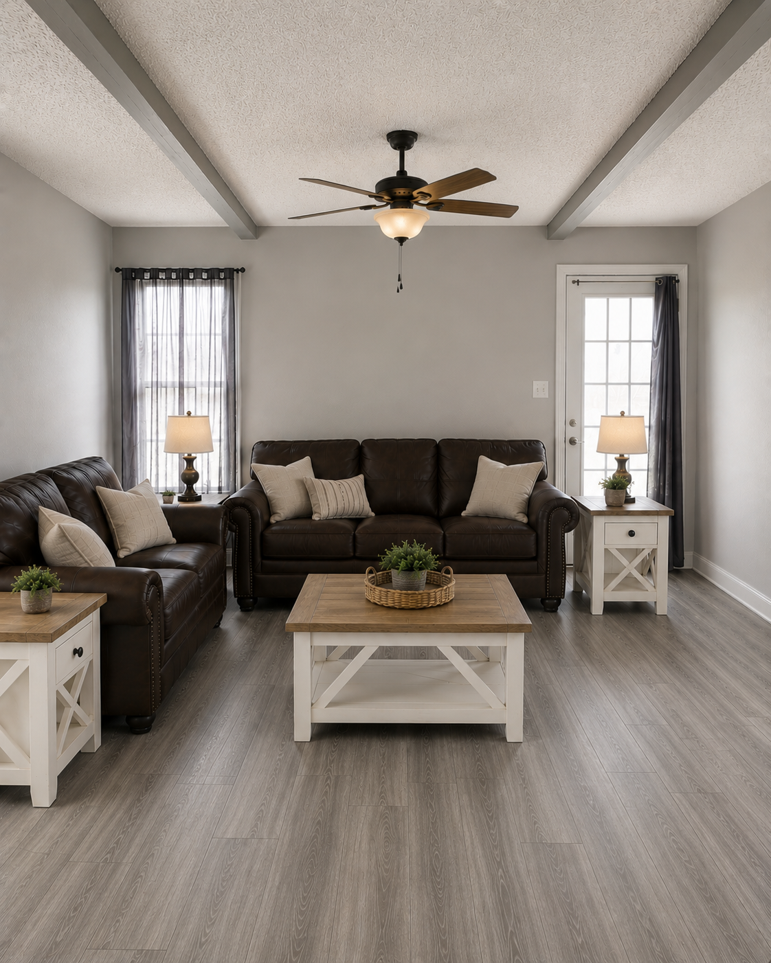 Living room with beige walls, gray ceiling beams, a ceiling fan with a light, large windows with dark curtains, a central dark brown leather sofa with beige cushions, two matching side tables with lamps, and two white coffee tables with plants.