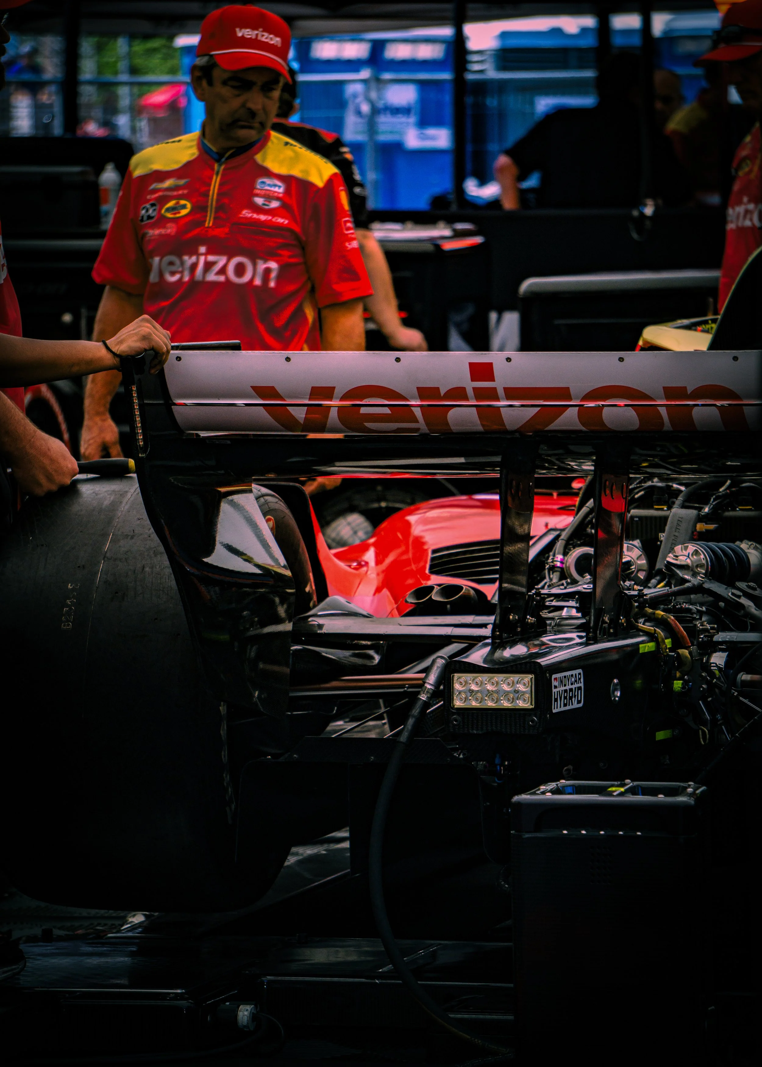 Race car with open engine and crew members working on it in the garage, person wearing a red Verizon racing shirt and hat.