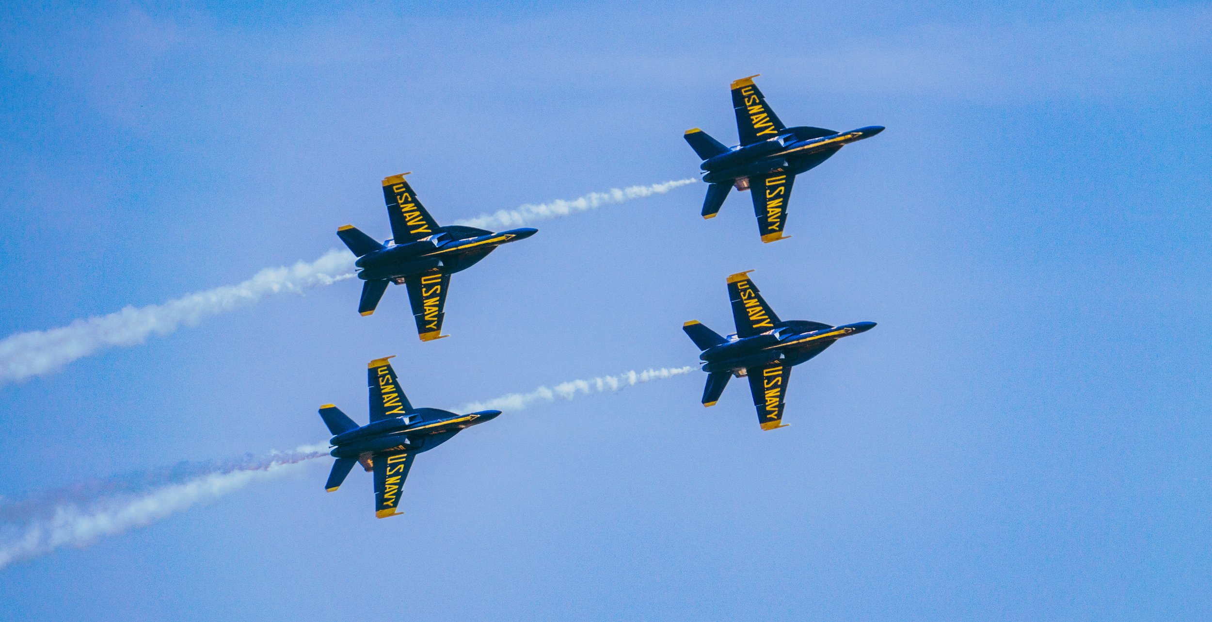 Four US Navy jet planes flying in a tight formation through the sky, leaving white smoke trails behind them.