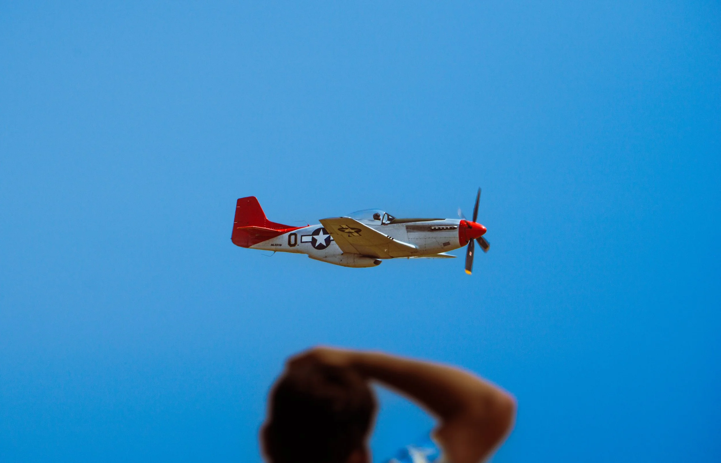 A person holding a camera and taking a picture of a vintage airplane flying in a clear blue sky.