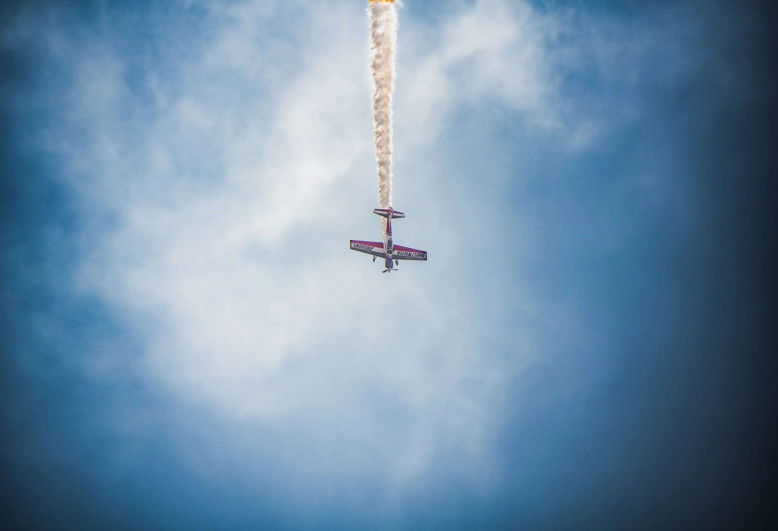 A small airplane performing an aerobatic stunt in the sky, leaving a trail of white smoke behind it. The sky is blue with some clouds.