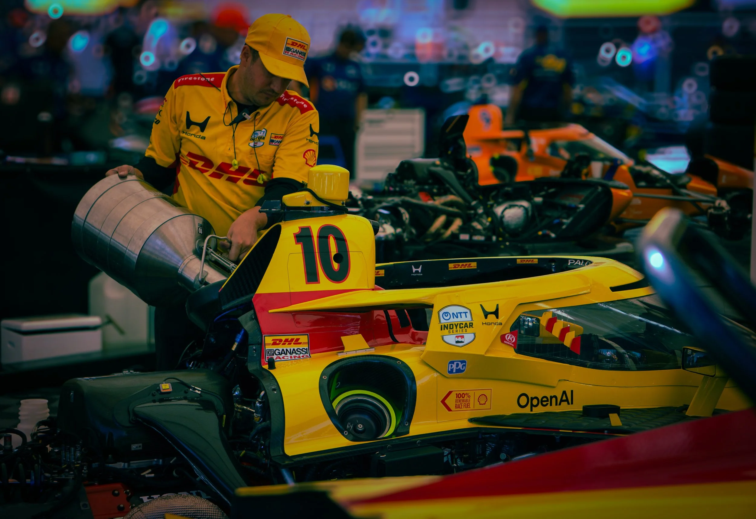 A man in a yellow racing suit and matching cap, working on a yellow Formula 1 race car in a garage with other racing cars and equipment in the background.