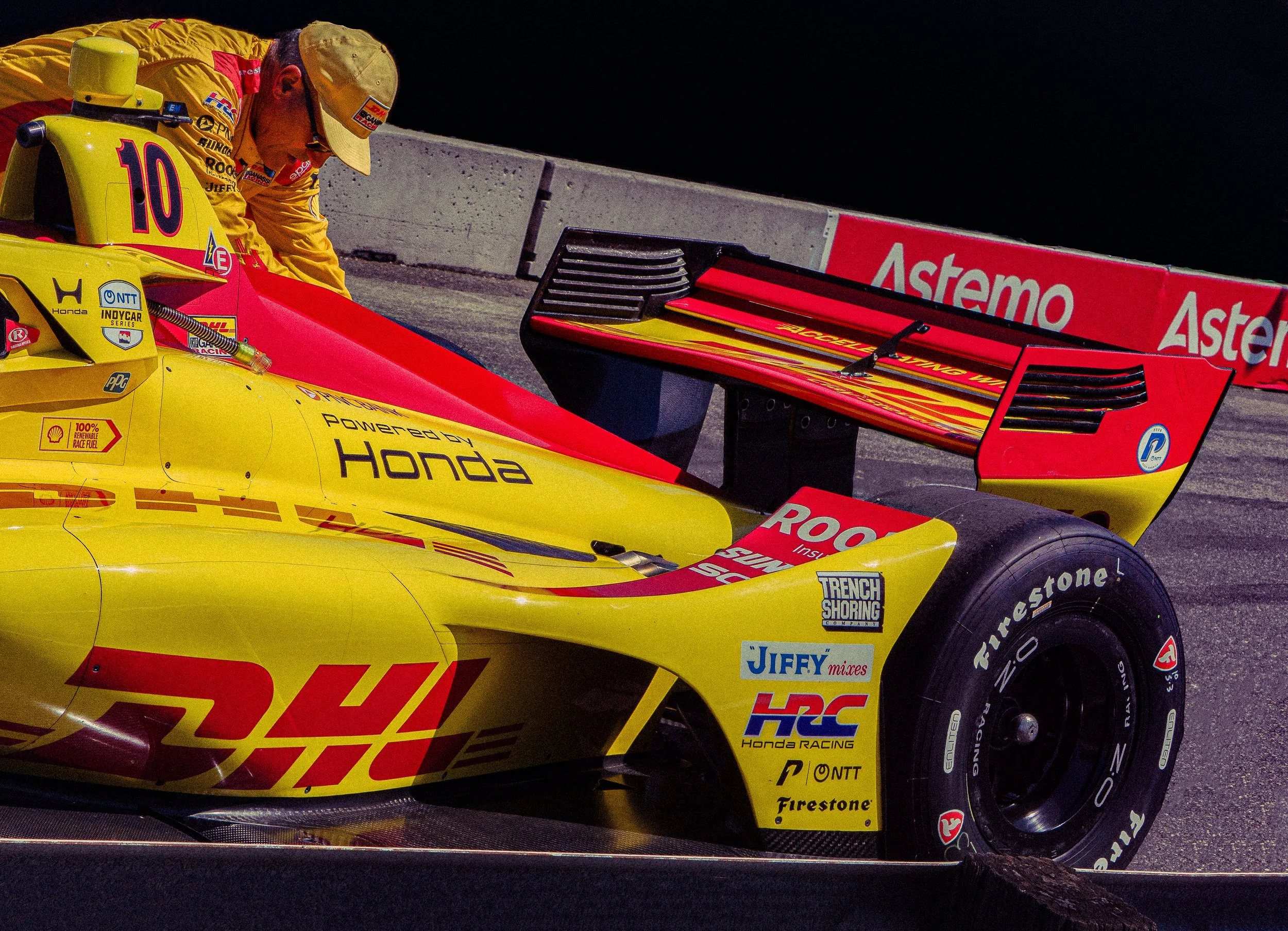 A race car driver in yellow racing gear working on a yellow and red IndyCar with various sponsor logos, including DHL, Honda, and Firestone, on a race track at night.