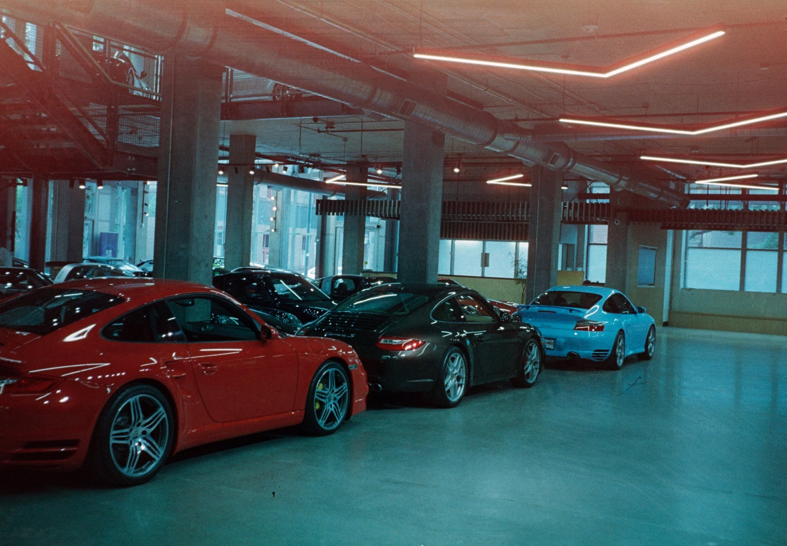 Luxury sports cars parked inside a modern parking garage with high ceilings, exposed ductwork, and illuminated ceiling lines.