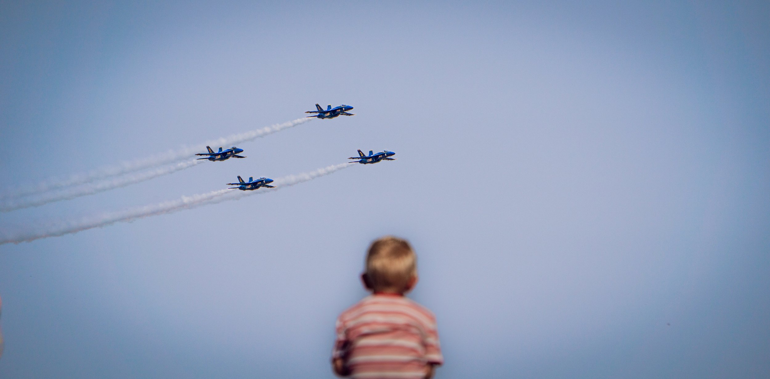 A young child with short hair and wearing a striped shirt watches four blue fighter jets flying in formation against a clear blue sky, leaving white vapor trails.