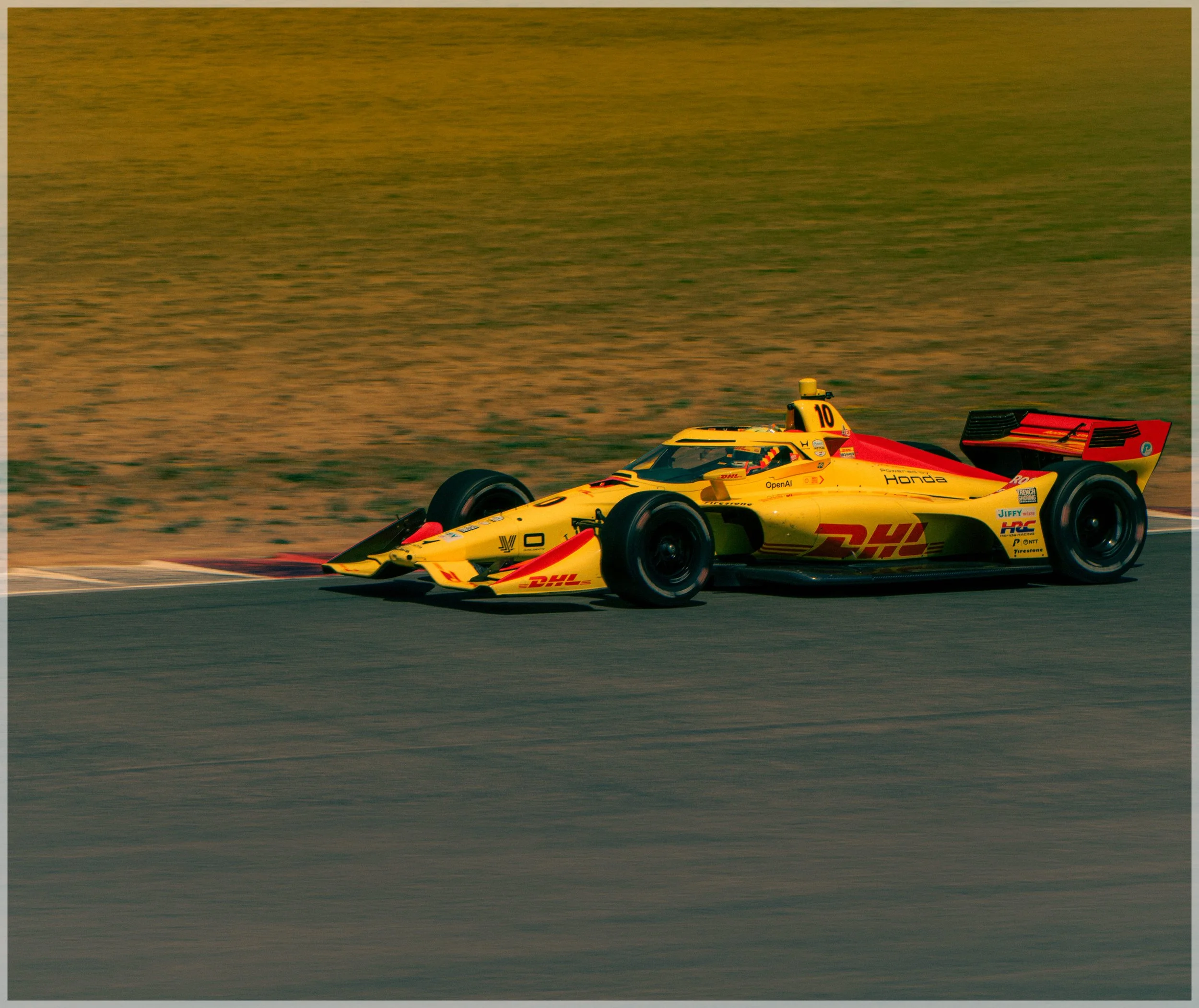 A yellow race car with sponsor logos and the Honda logo on the side, speeding on a track during sunset with a blurred background.