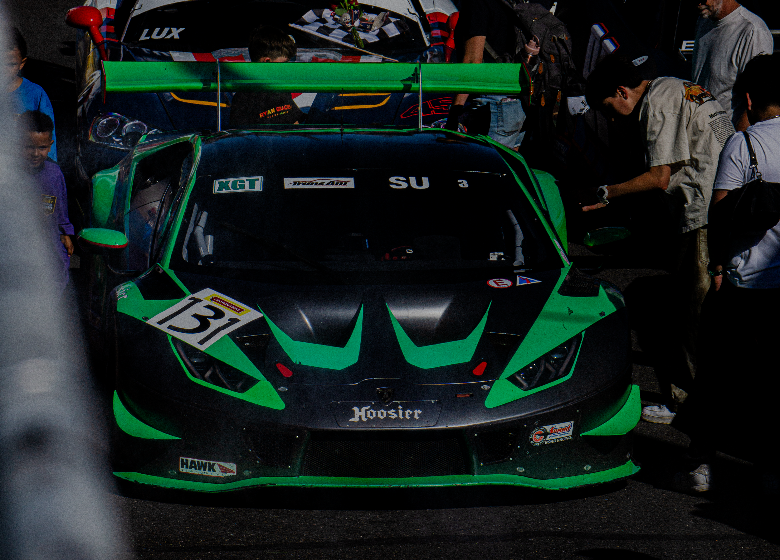 A group of people gathered around a green and black race car at a motorsport event. The car has stickers, a large rear spoiler, and visible racing decals. Some individuals are inspecting or preparing the car.