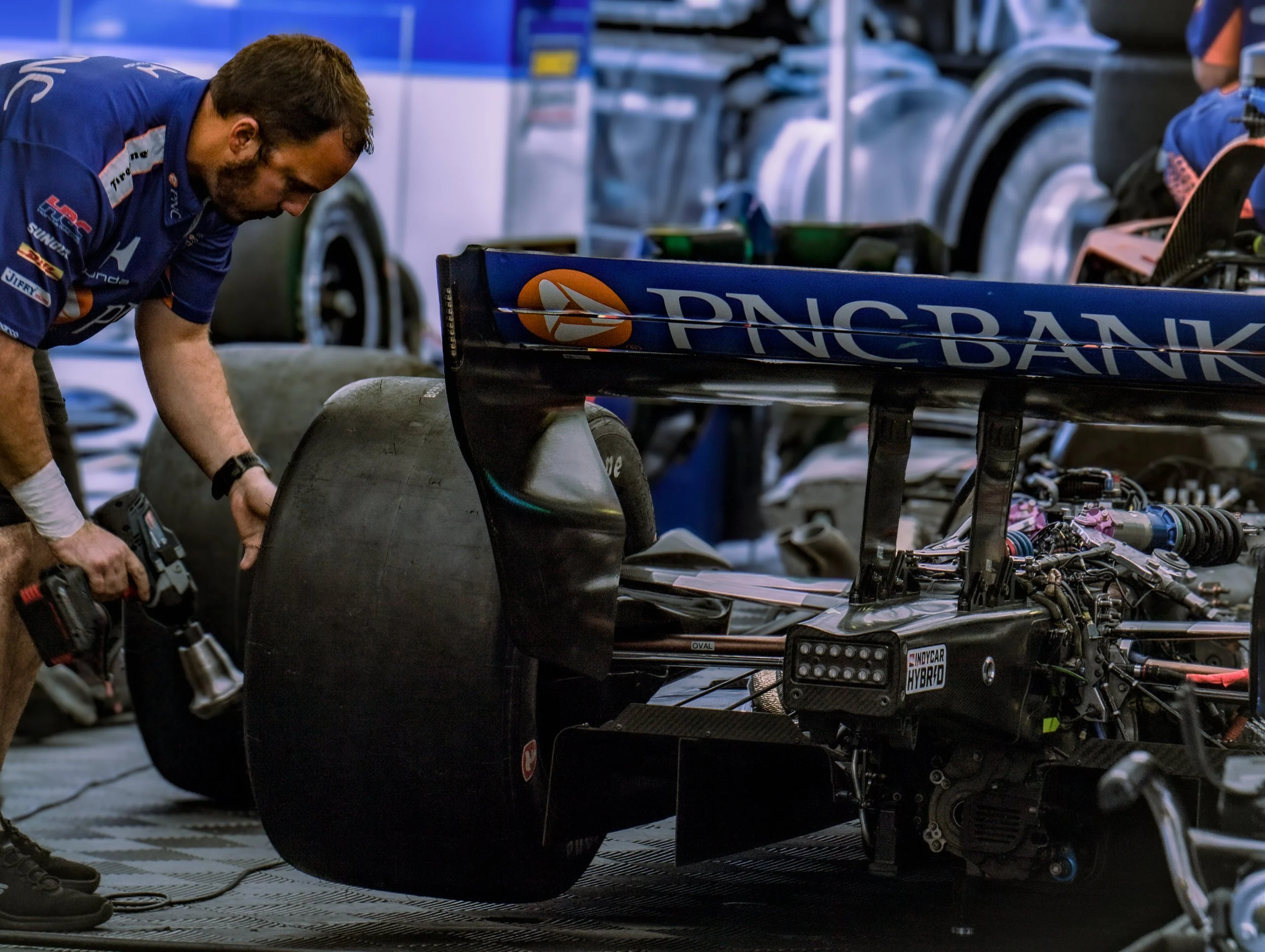 A mechanic working on a Formula 1 race car in the garage, focusing on the rear wing with brands like PNC Bank visible, and various parts and tools around.