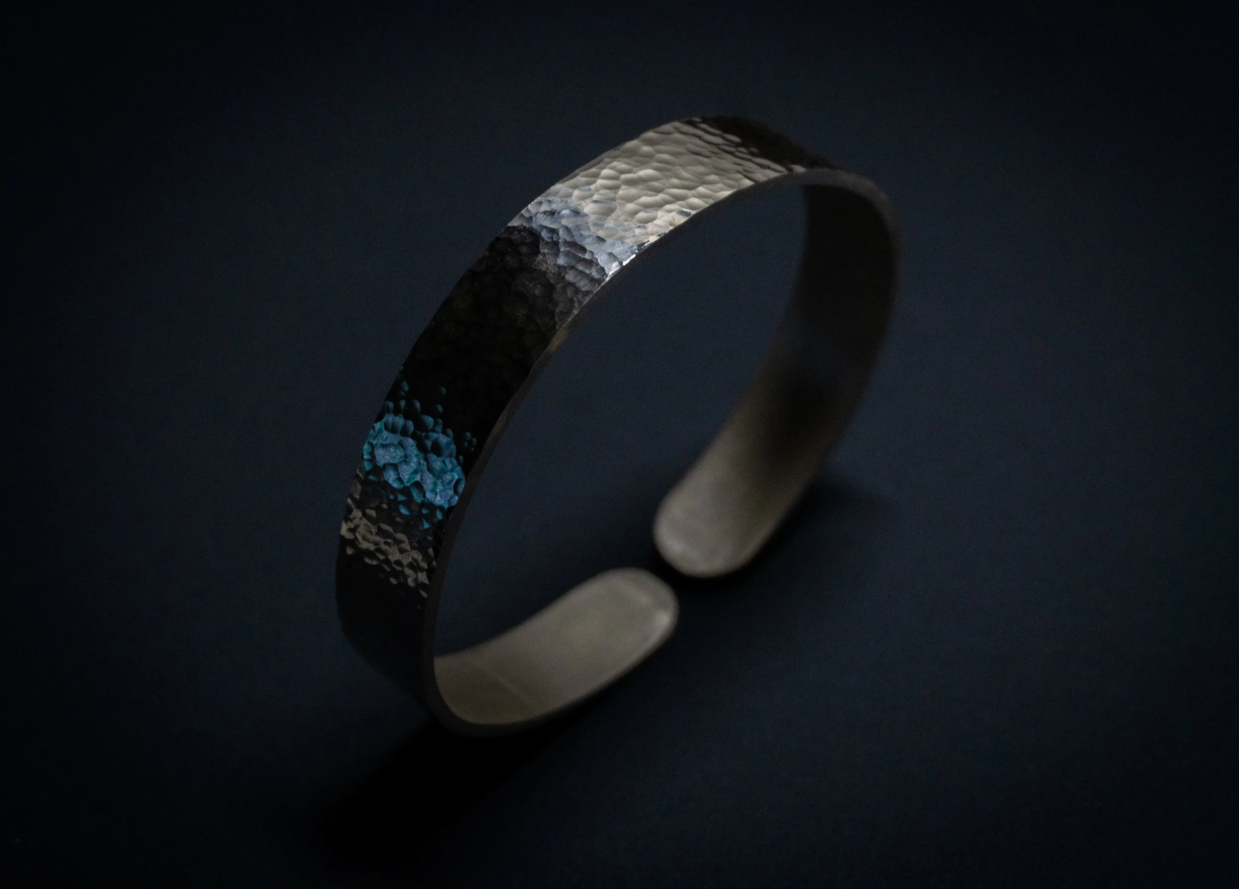Close-up of a hammered metal band ring with textured surface and reflective finish, set against a dark background.
