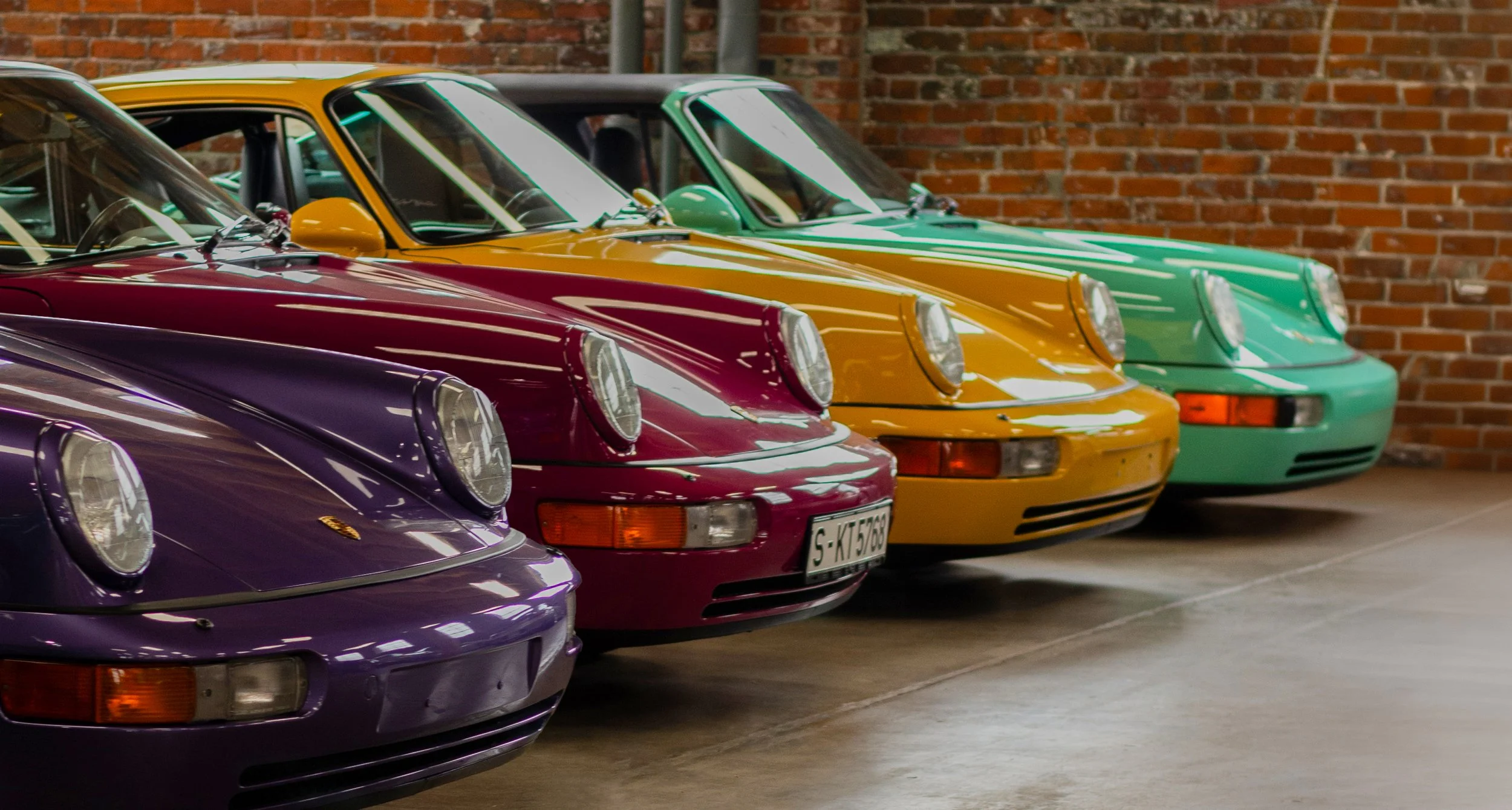 A row of colorful vintage Porsche 911 cars parked inside a garage with a brick wall backdrop.