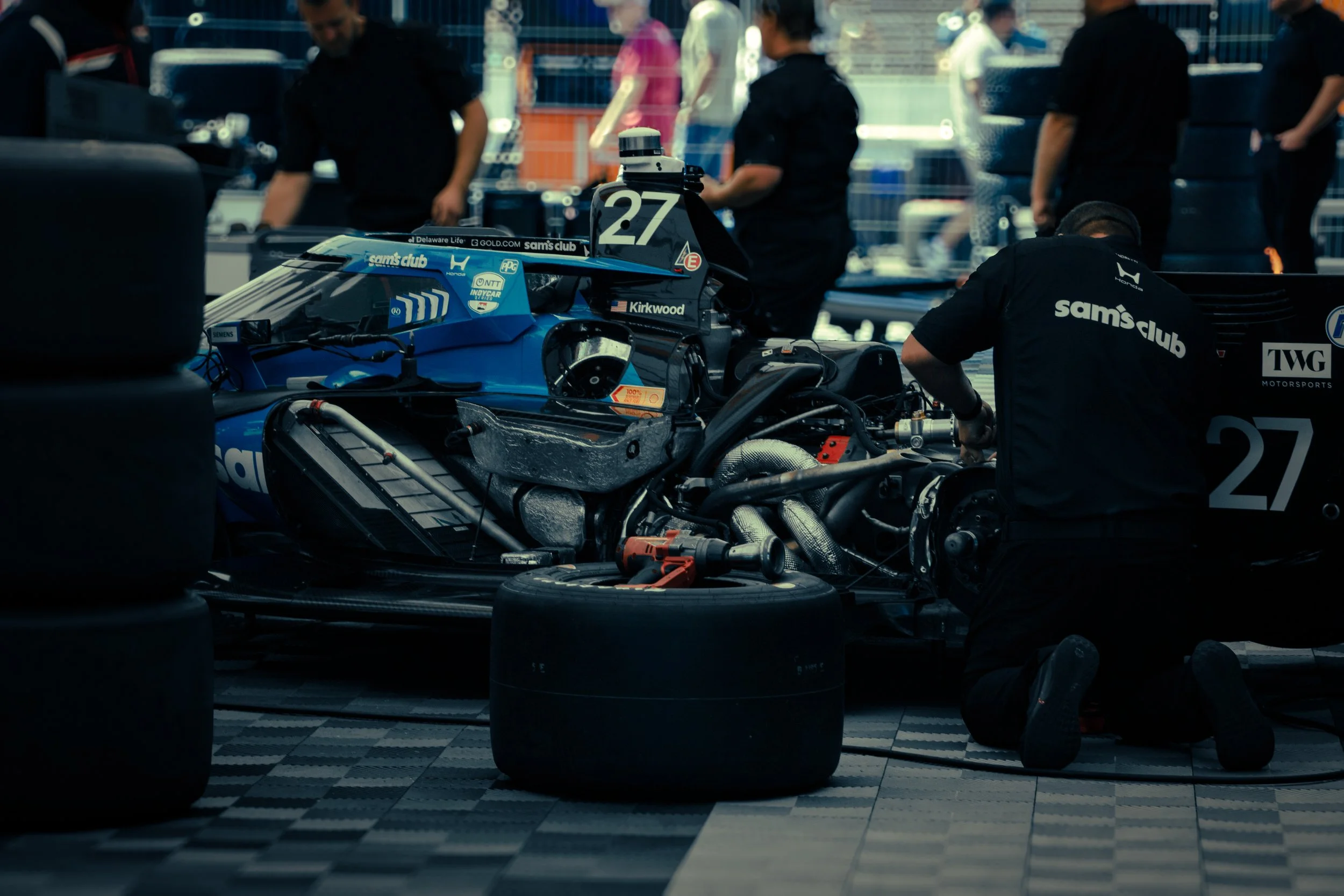 A race car in a garage during maintenance with team members working on it, surrounded by tires and equipment.