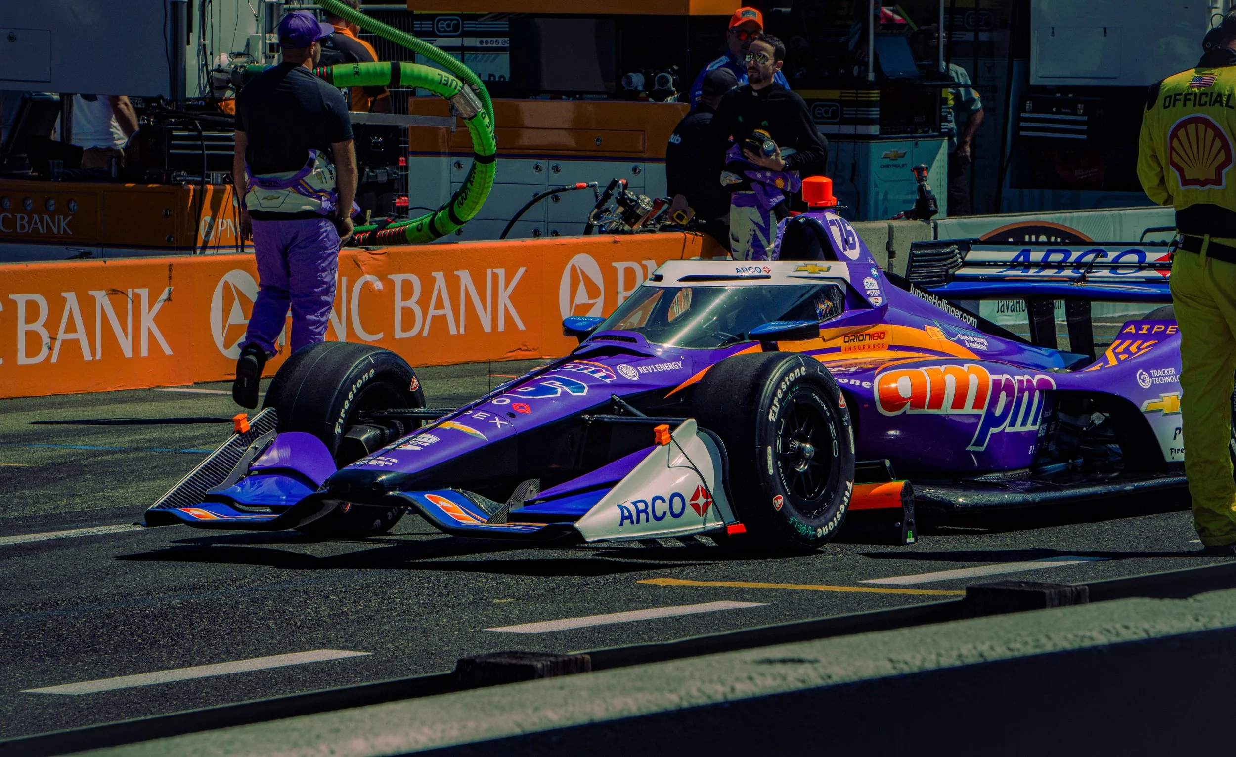 Race car in the pit lane with crew members working around it. The car is purple with sponsor logos, and there are various equipment and personnel preparing for a race.