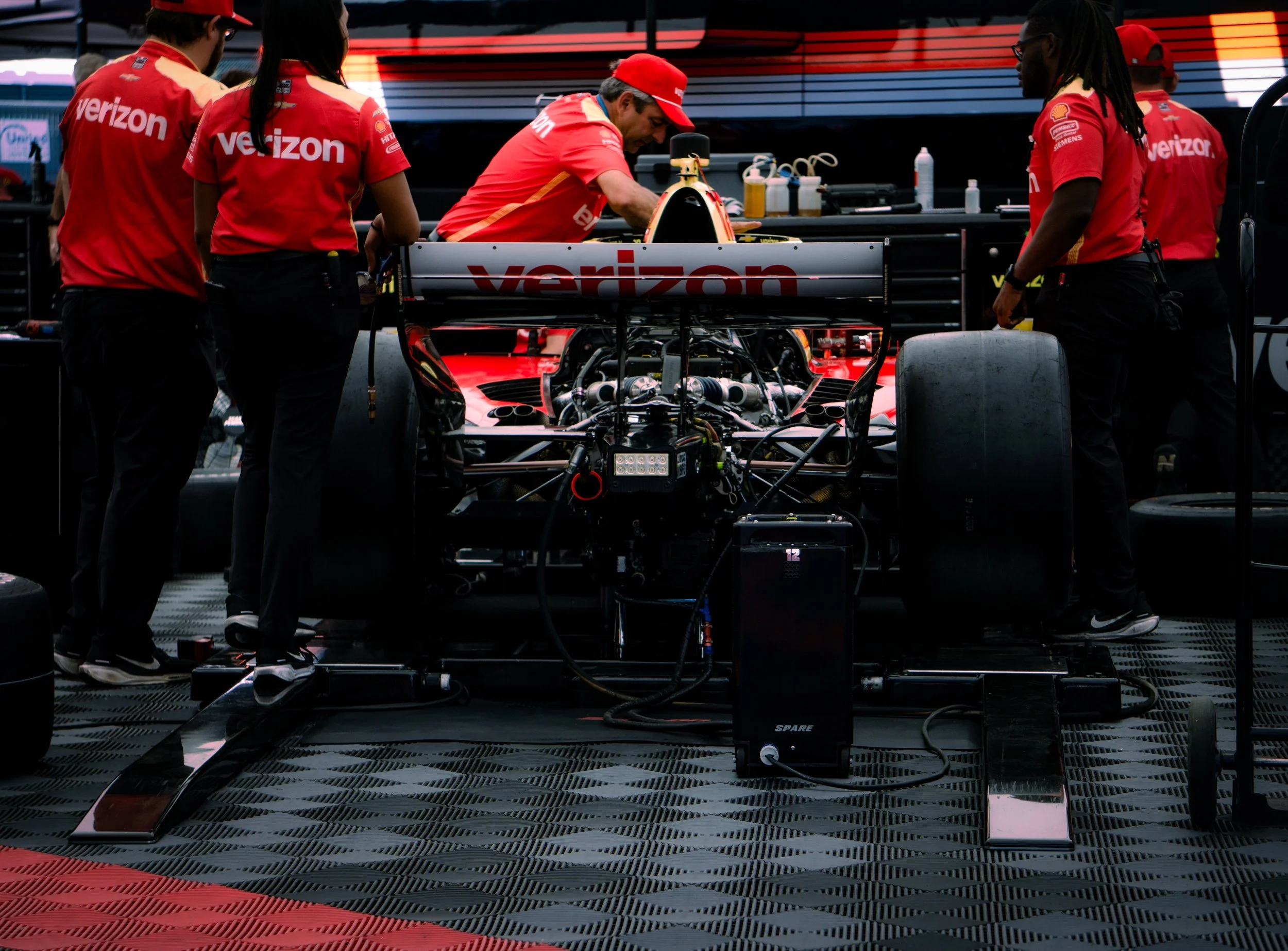 Race car team members working on a Formula 1 car in the garage, with team uniforms displaying the Verizon logo.