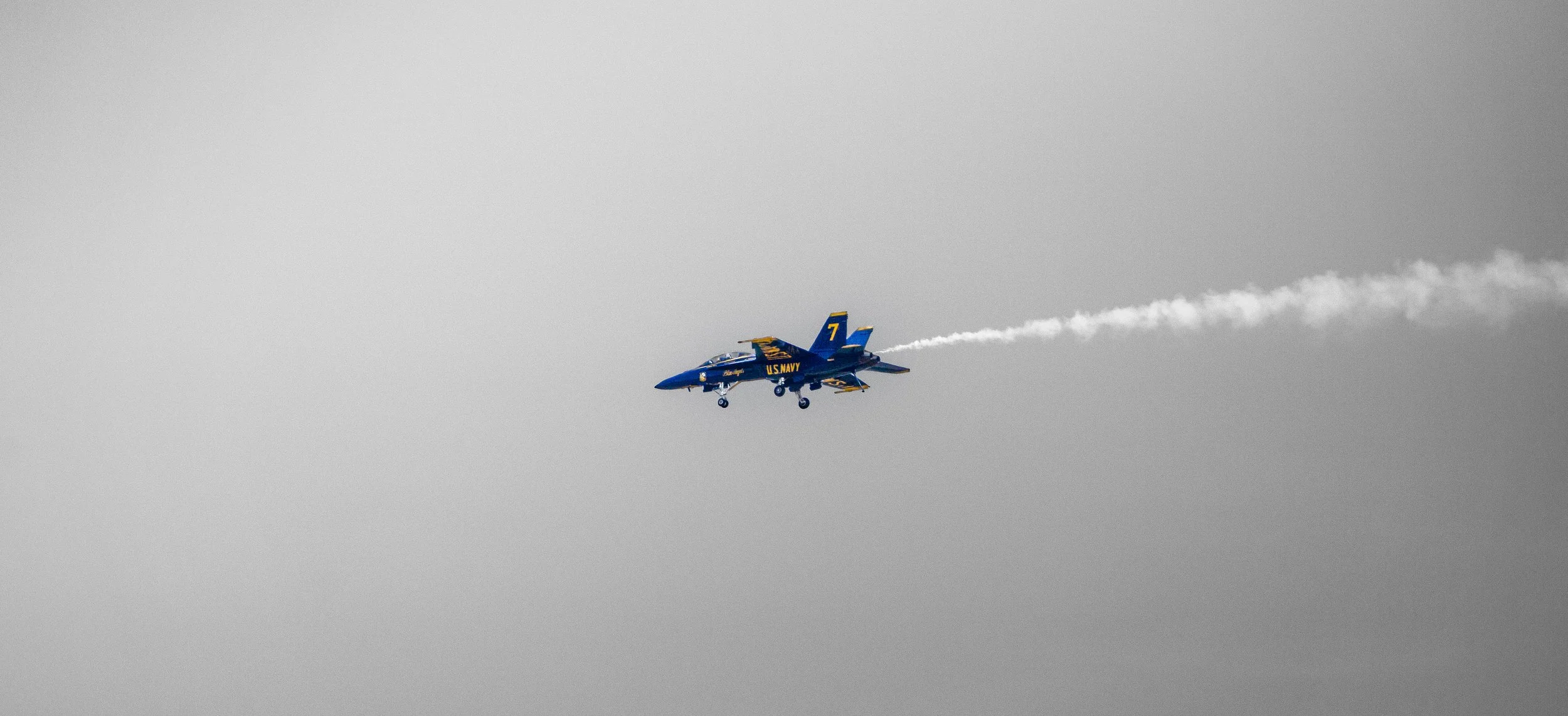 A blue and yellow U.S. Navy fighter jet flying through the gray sky, leaving a white smoke trail behind.