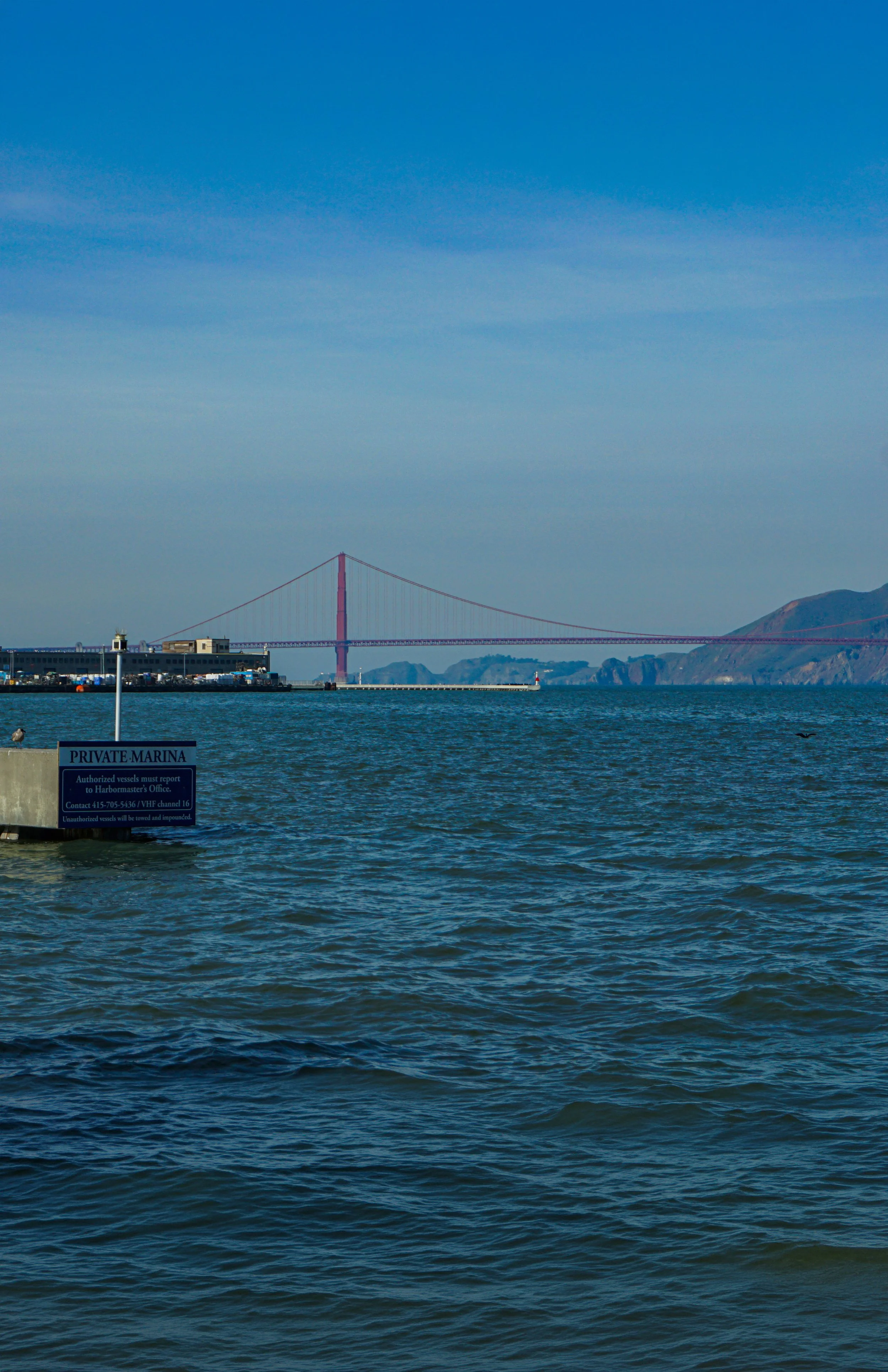 View of the bay with the Golden Gate Bridge in the distance and a marina in the foreground. The sky is blue with light clouds.
