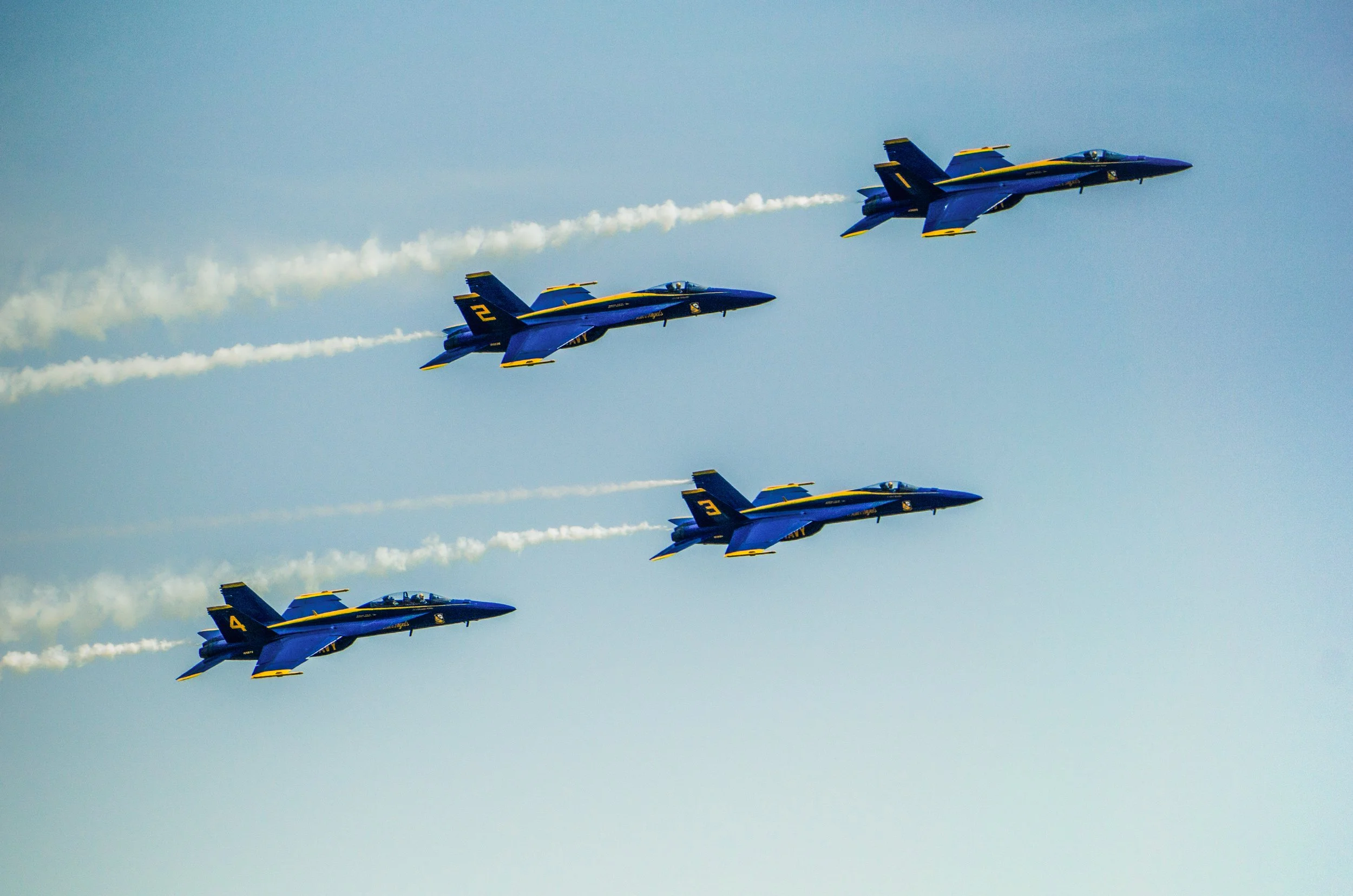 Group of blue fighter jets flying in formation, emitting white smoke trails against a clear blue sky.