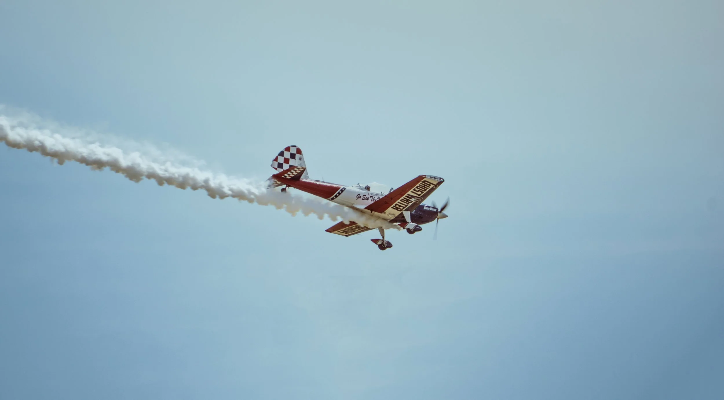 An aerobatic airplane with red and white checkered tail, flying upside down with smoke trailing behind against a blue sky.
