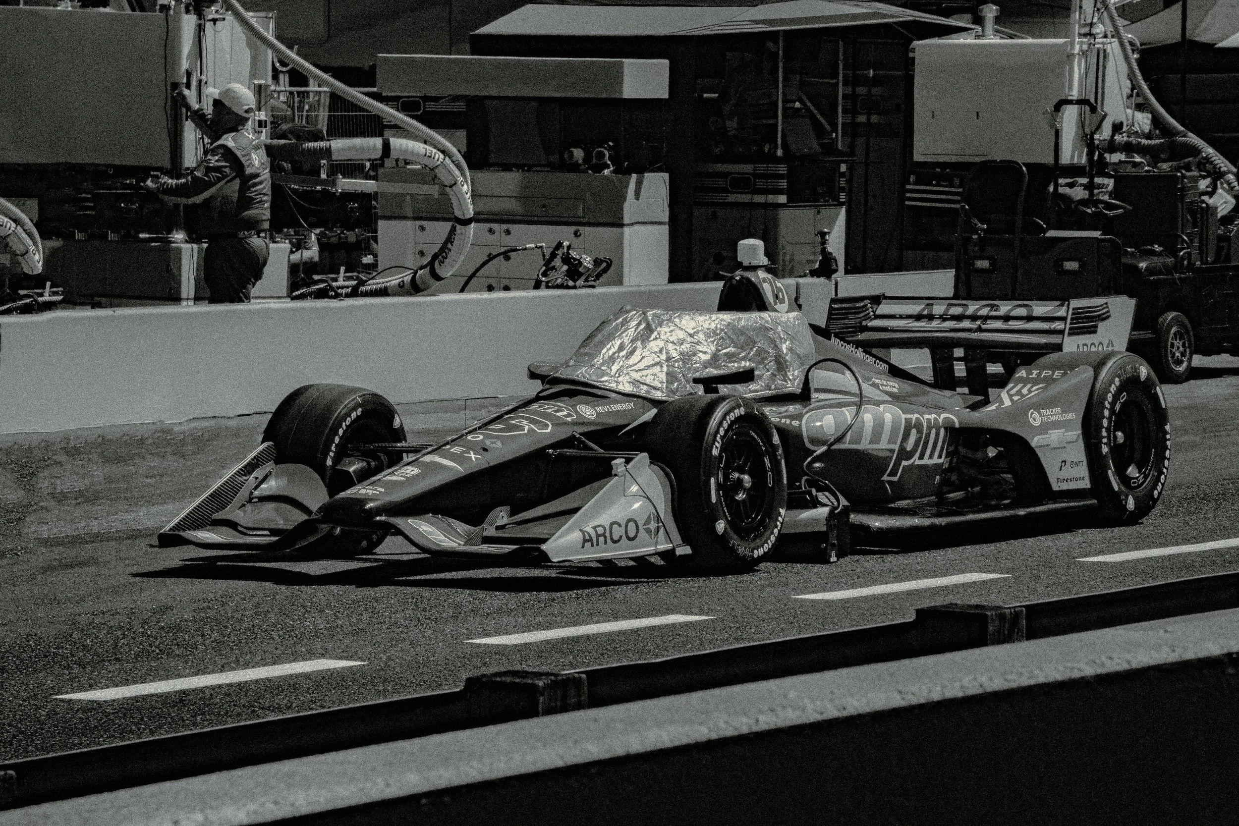 A race car parked in the pit lane at night, with a mechanic working on or inspecting it. The car has sponsorship logos and a streamlined design.