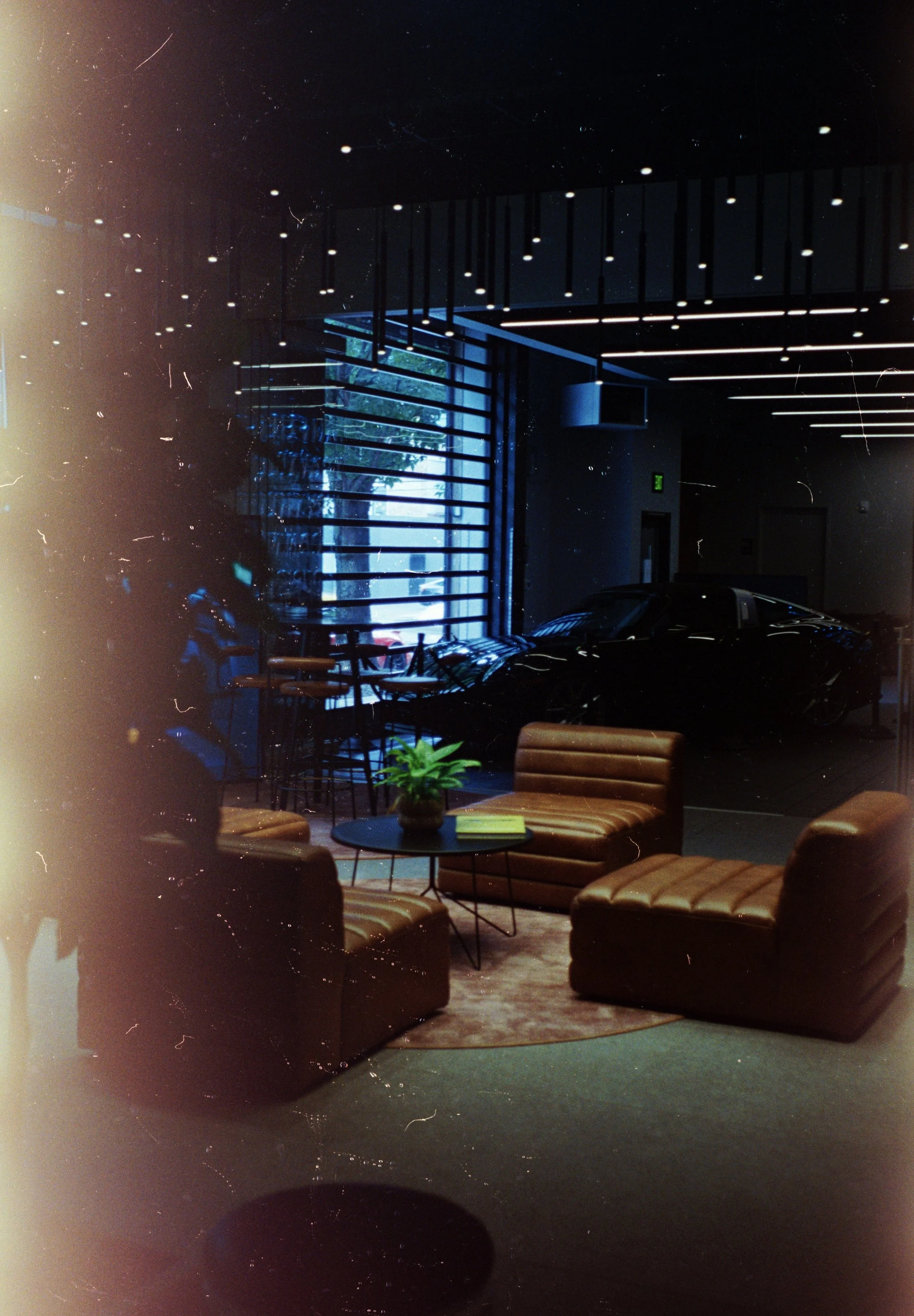 Interior of a modern lounge or waiting area with leather chairs, a small coffee table with a potted plant and books, large windows with blue lighting, ceiling with hanging lights, and a dark-colored interior.