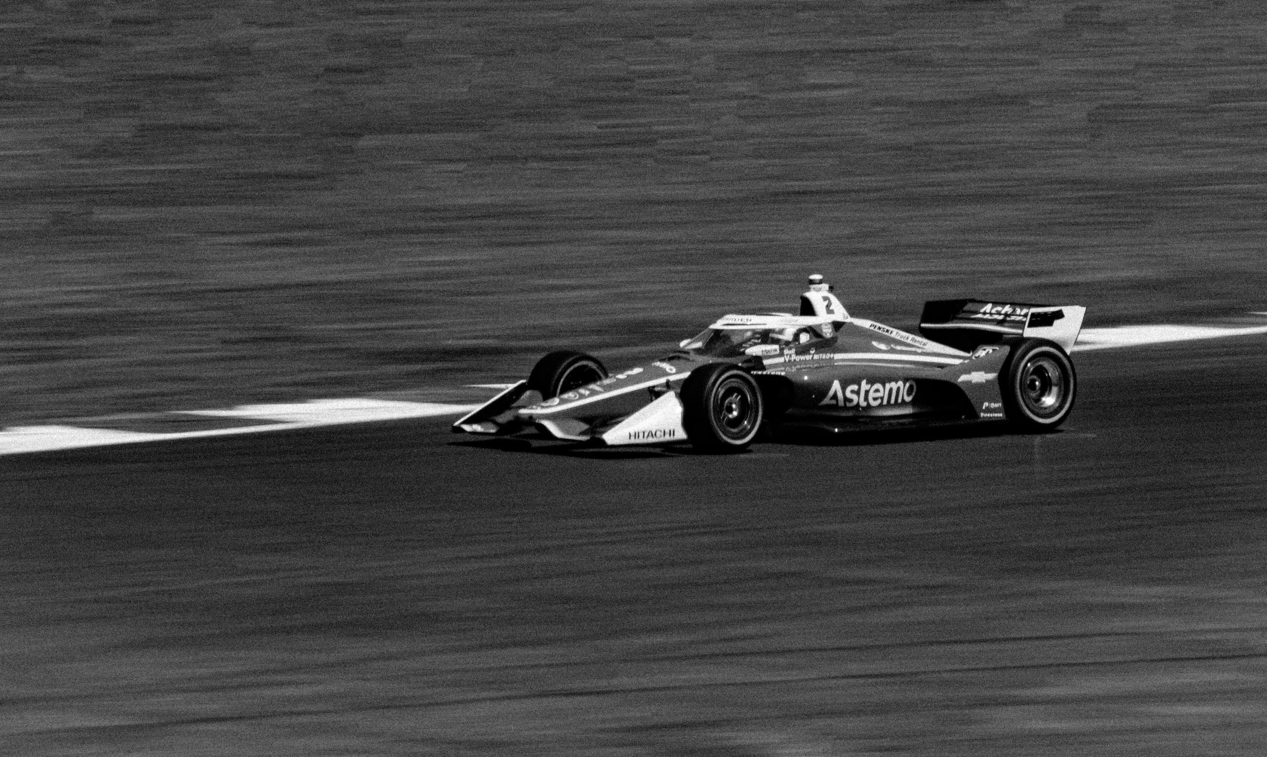 A race car on a track in black and white, with sponsorship markings including Astemo and Hitachi, driven by a driver wearing a helmet, speeding along the curve of the racetrack.