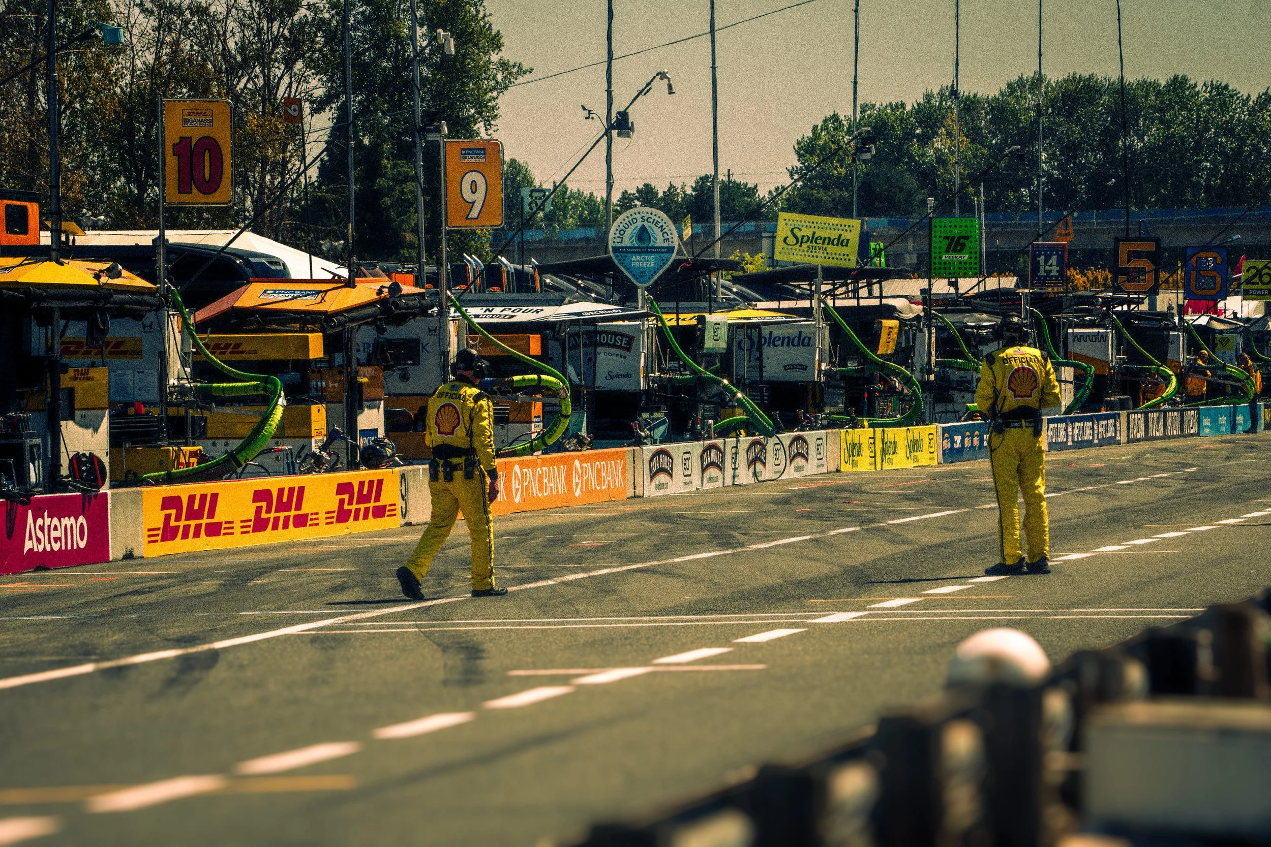 Race track with a row of race cars and team personnel wearing yellow Shell suits, with banners and logos advertising DHL, Splenda, and other sponsors, and numbered signs indicating different racing positions.