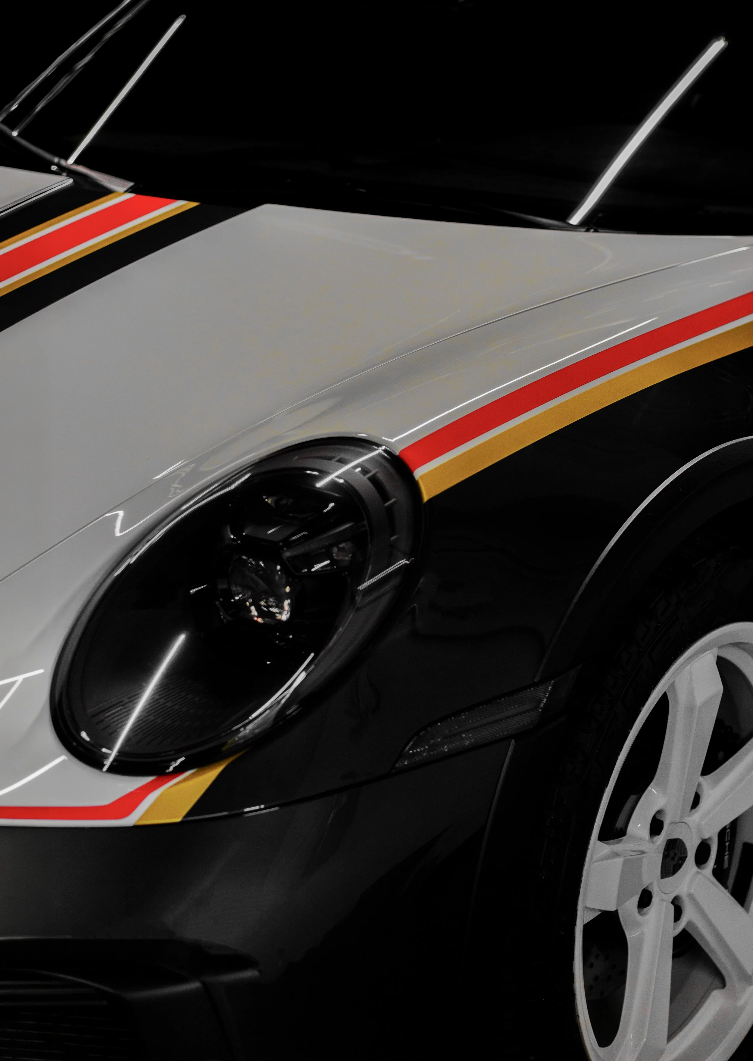 Close-up view of a sports car with a white body, black roof, and racing stripes in red, orange, and yellow, featuring a silver alloy wheel.