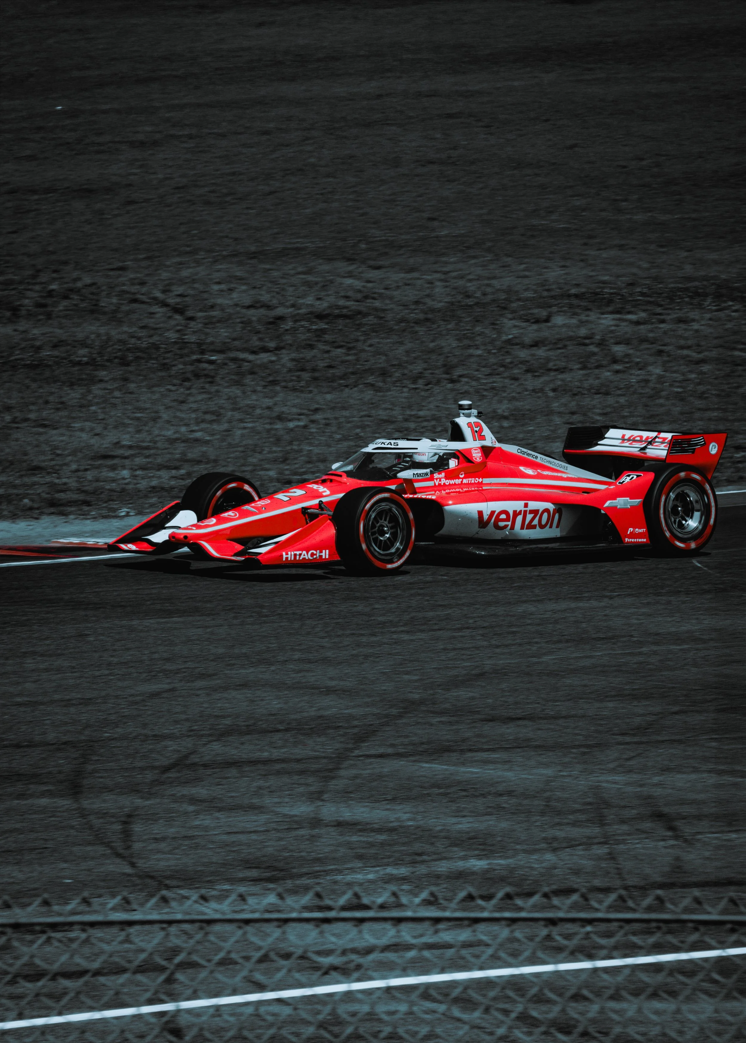 A red and white Verizon-sponsored IndyCar race car on a dark race track, viewed from the side with a chain-link fence in the foreground.