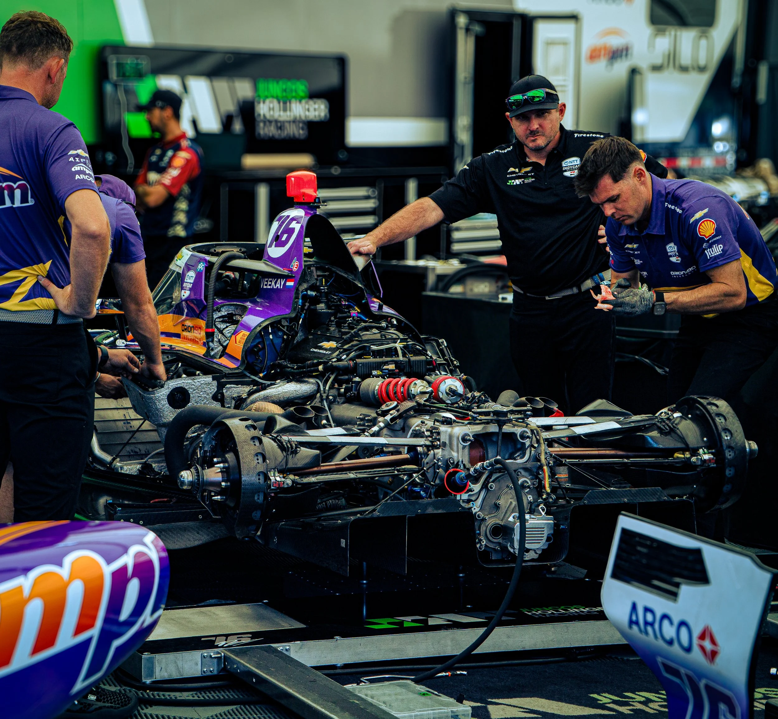 A group of mechanics working on a purple race car in a racing garage, with tools and equipment around.