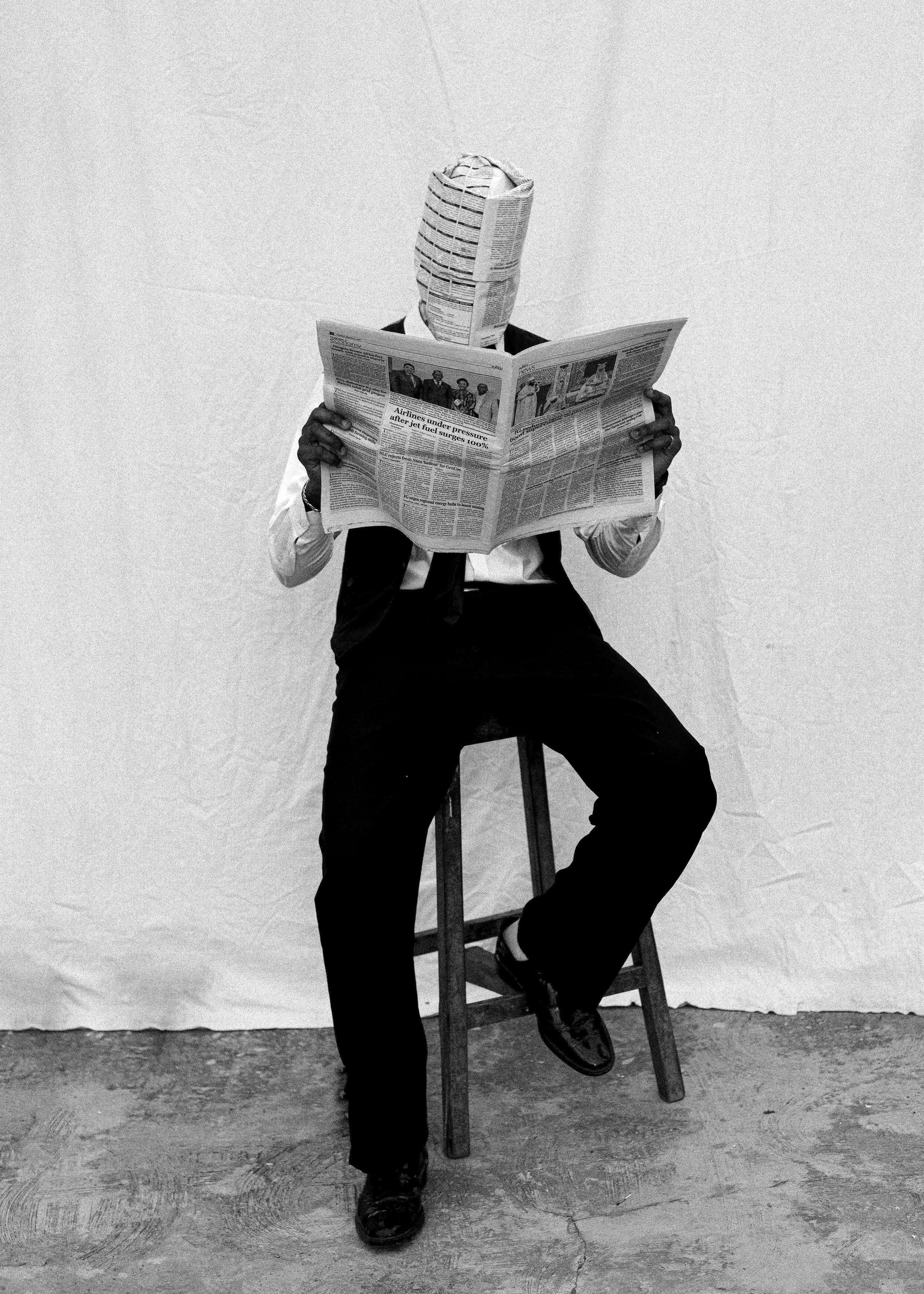 Person sitting on a wooden chair holding a newspaper, with their face covered by a crumpled newspaper, in front of a plain wall.