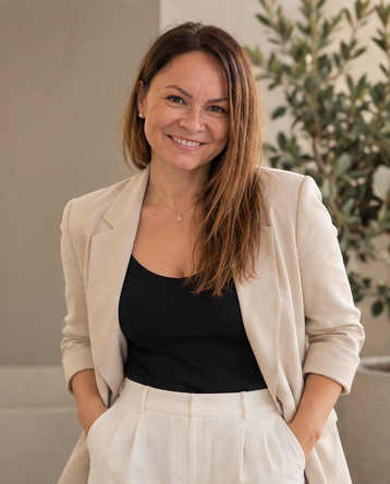 A woman with shoulder-length brown hair and a smile, standing indoors with a plant in the background, wearing a beige blazer over a black top.