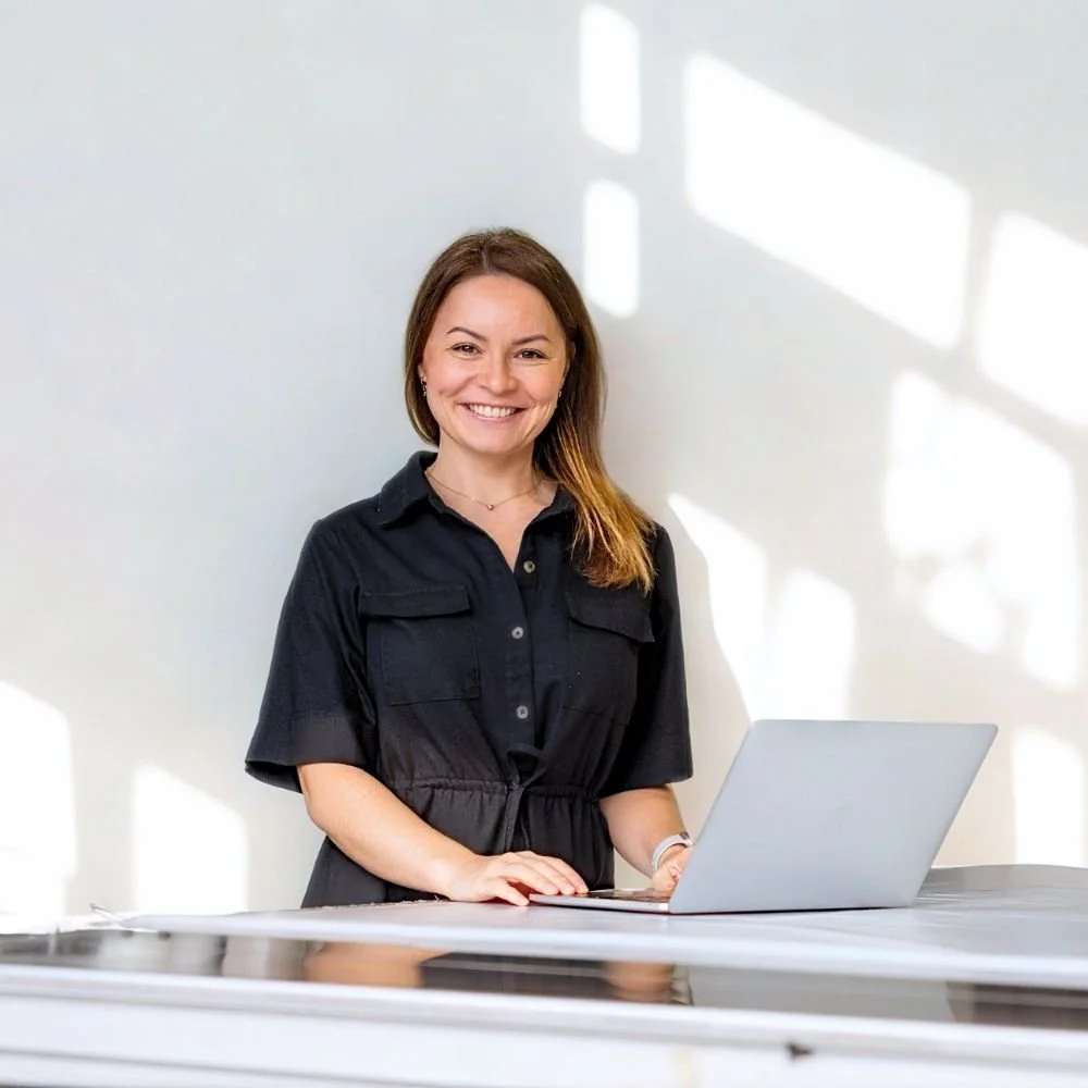 A woman with brown hair smiling while standing at a table with a laptop in front of her, in a room with sunlight creating shadows on the wall.