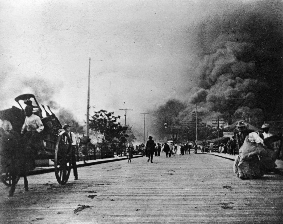 Black-and-white historical photograph of Jacksonville, Florida, during the Great Fire of 1901, showing people and horse-drawn carts on a city street as thick smoke rises from burning buildings in the distance.