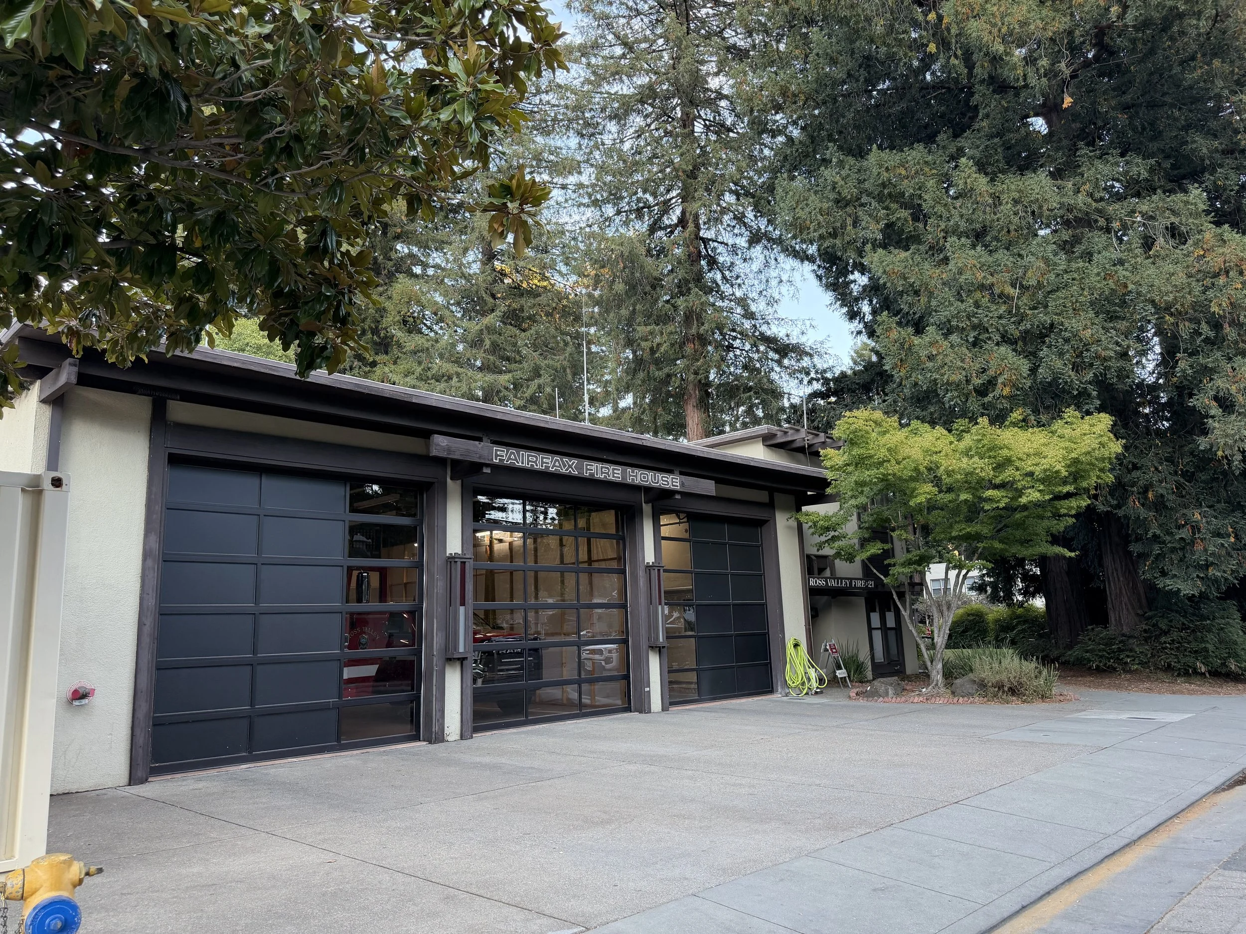 Fire station with a garage door, surrounded by large trees and a paved area in front.