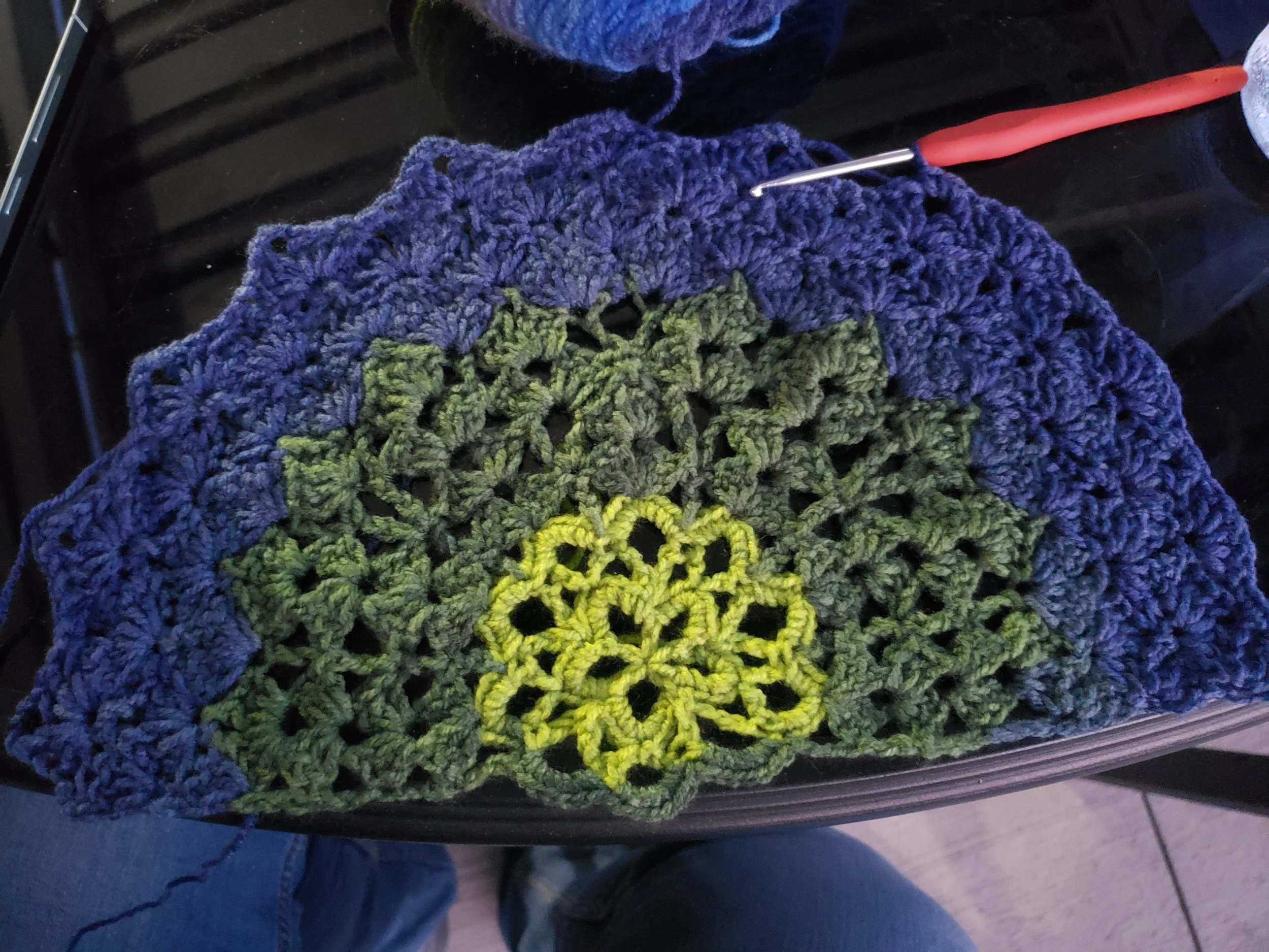 A crochet project made with multicolored yarn, transitioning from purple at the top to green and yellow at the bottom, placed on a black surface with a crochet hook inserted into the yarn.