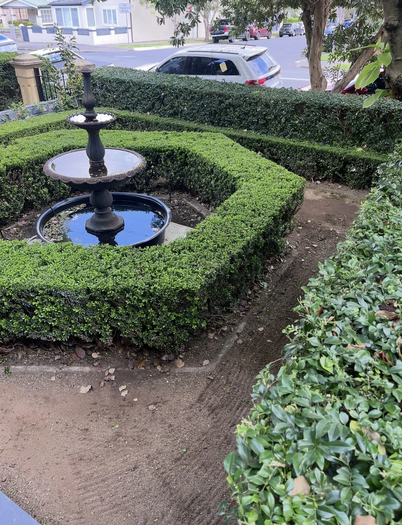 A small, decorative fountain with two tiers placed in the middle of a square hedge garden. The garden is surrounded by well-maintained bushes, and a walkway runs along its edge. In the background, a street with parked cars and trees is visible.