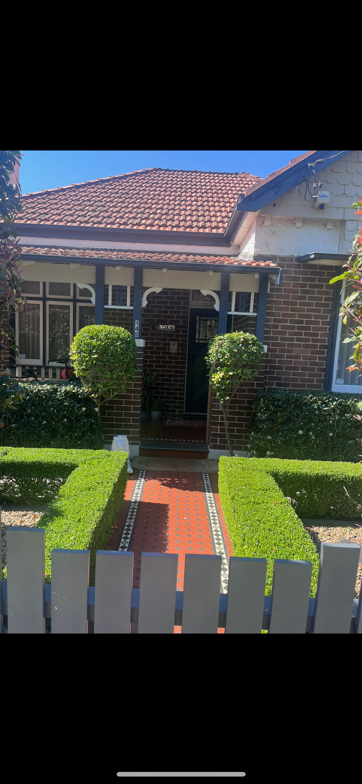 Front view of a brick house with a red tile roof, green bushes, and a decorative red and black tiled pathway leading to the front door. The house has a small porch with blue support beams and white trim.
