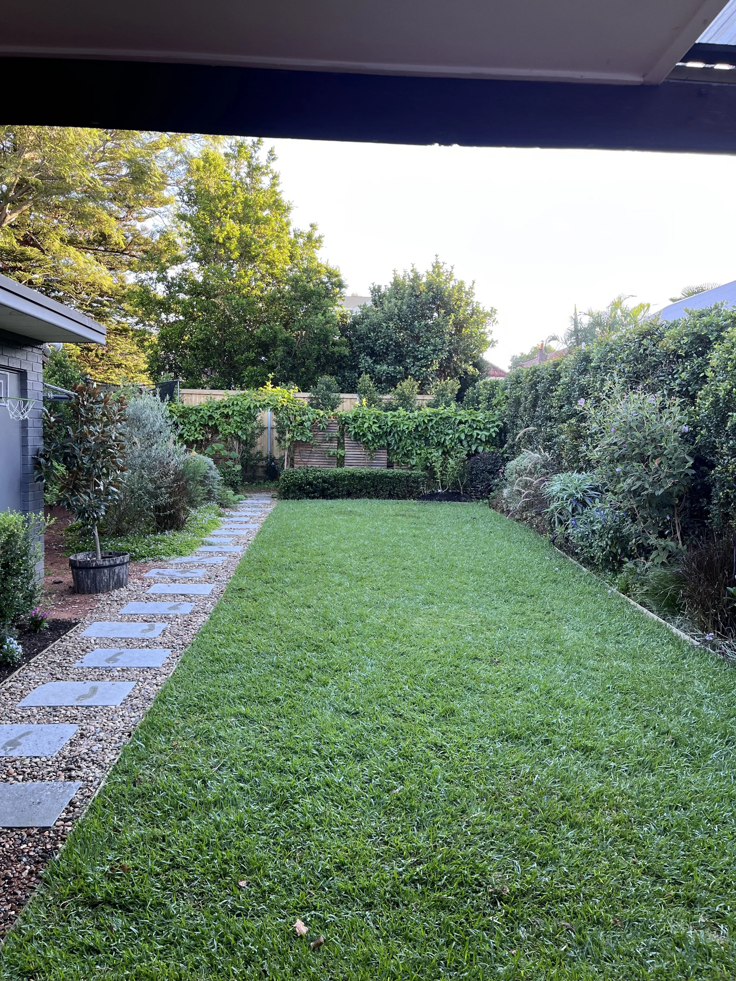 A well-maintained backyard with a green lawn, a stone and gravel pathway on the left, and various plants and bushes along the fence and house. Trees can be seen in the background with a clear sky overhead.