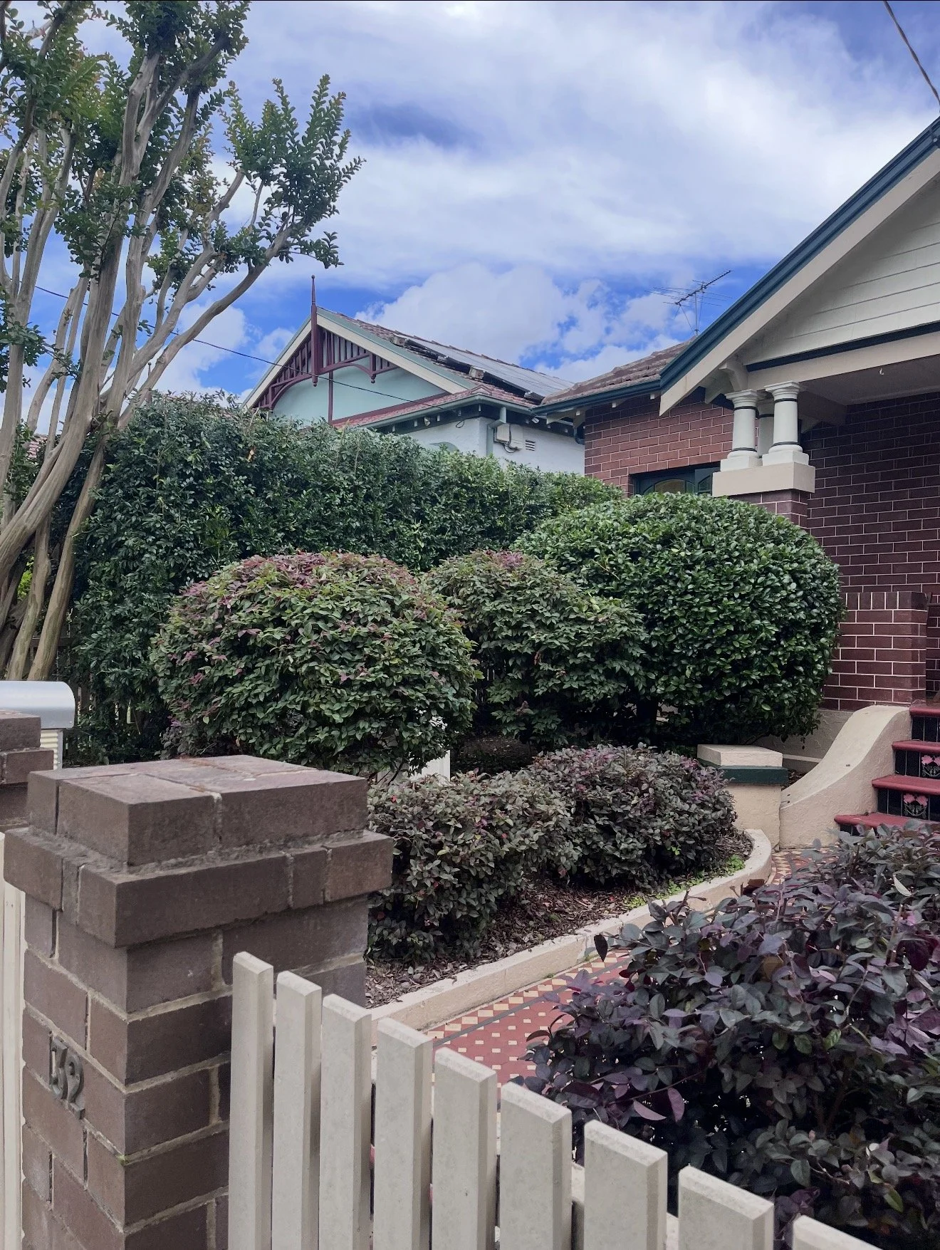 A front yard with a brick and white picket fence, landscaped bushes, a brick house with a covered porch, and a cloudy sky in the background.