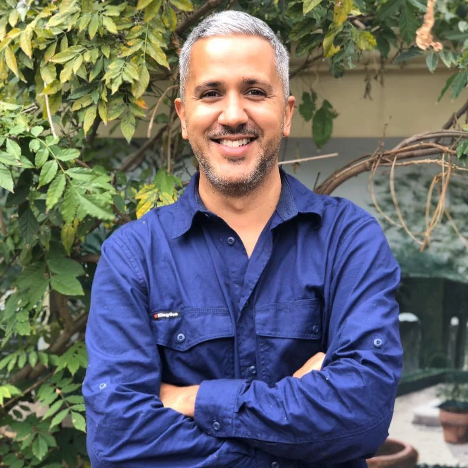 A smiling man with short gray hair and a beard, wearing a blue button-up shirt, standing outdoors in front of green leafy plants.
