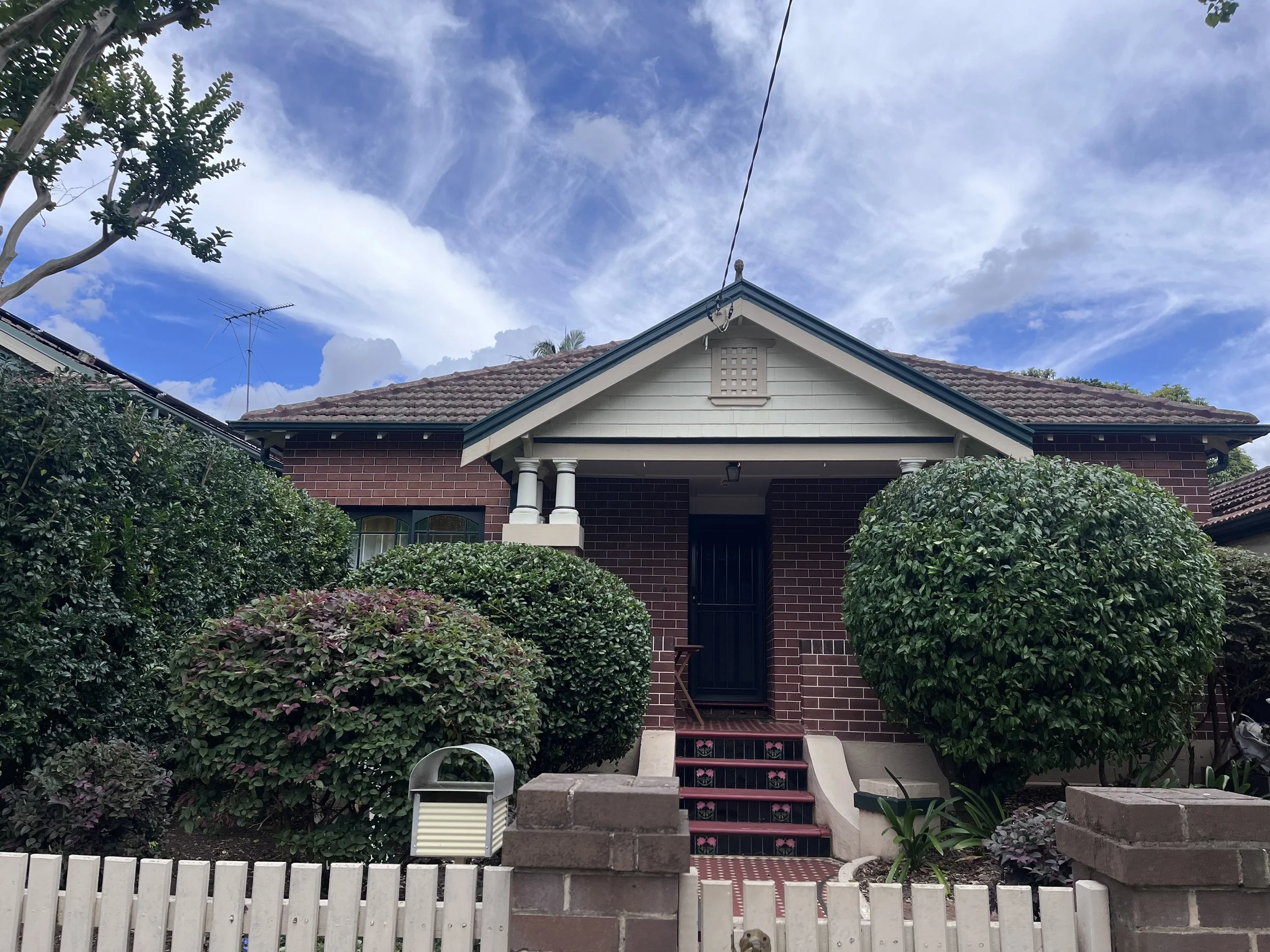 A house with a brick exterior, white columns at the entrance, a tiled roof, and lush green bushes in the front yard.