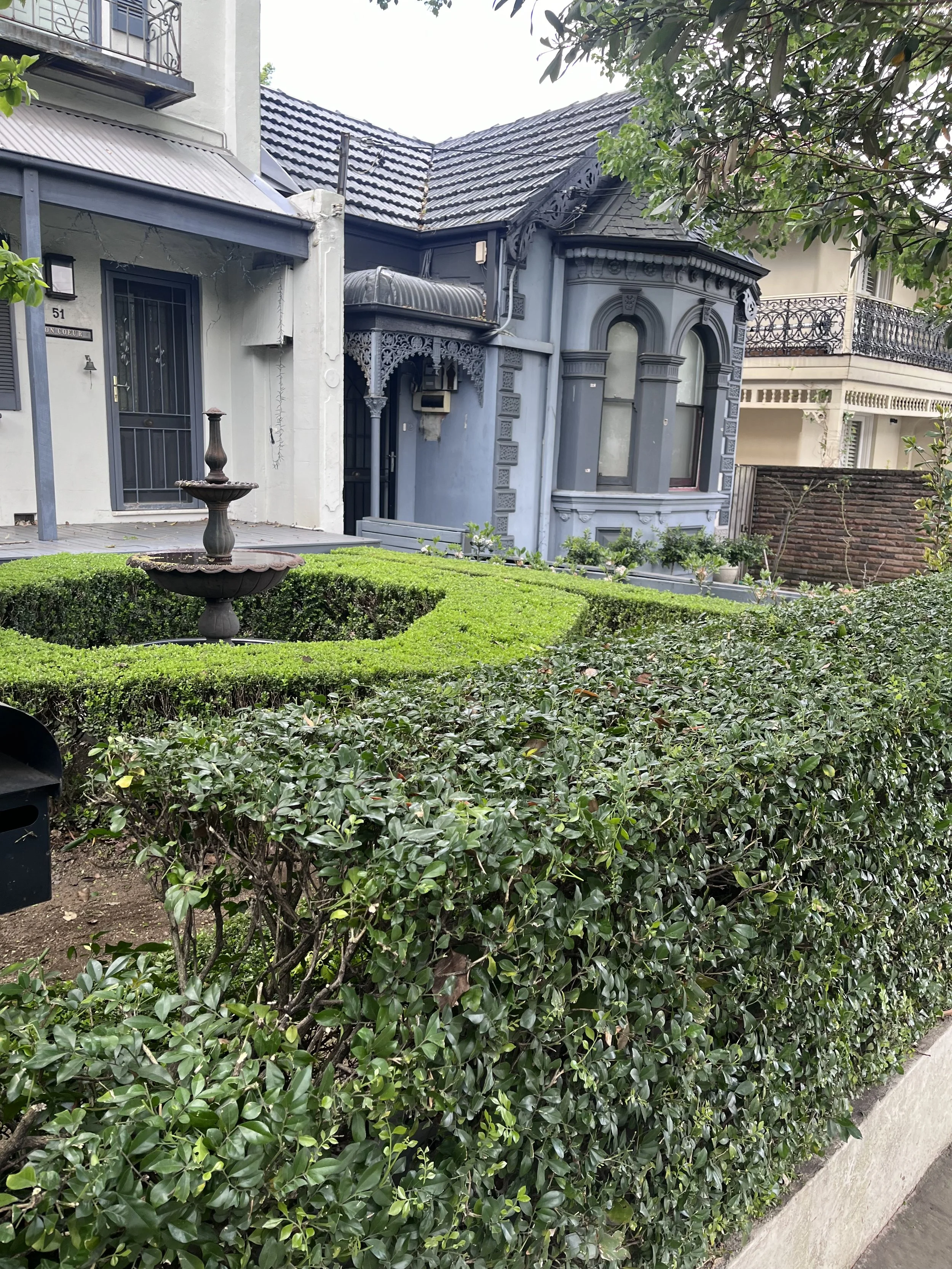 Front yard with neatly trimmed bushes, a decorative fountain, and a two-story house with a light-colored exterior, second-floor balcony, and a dark-colored roof.
