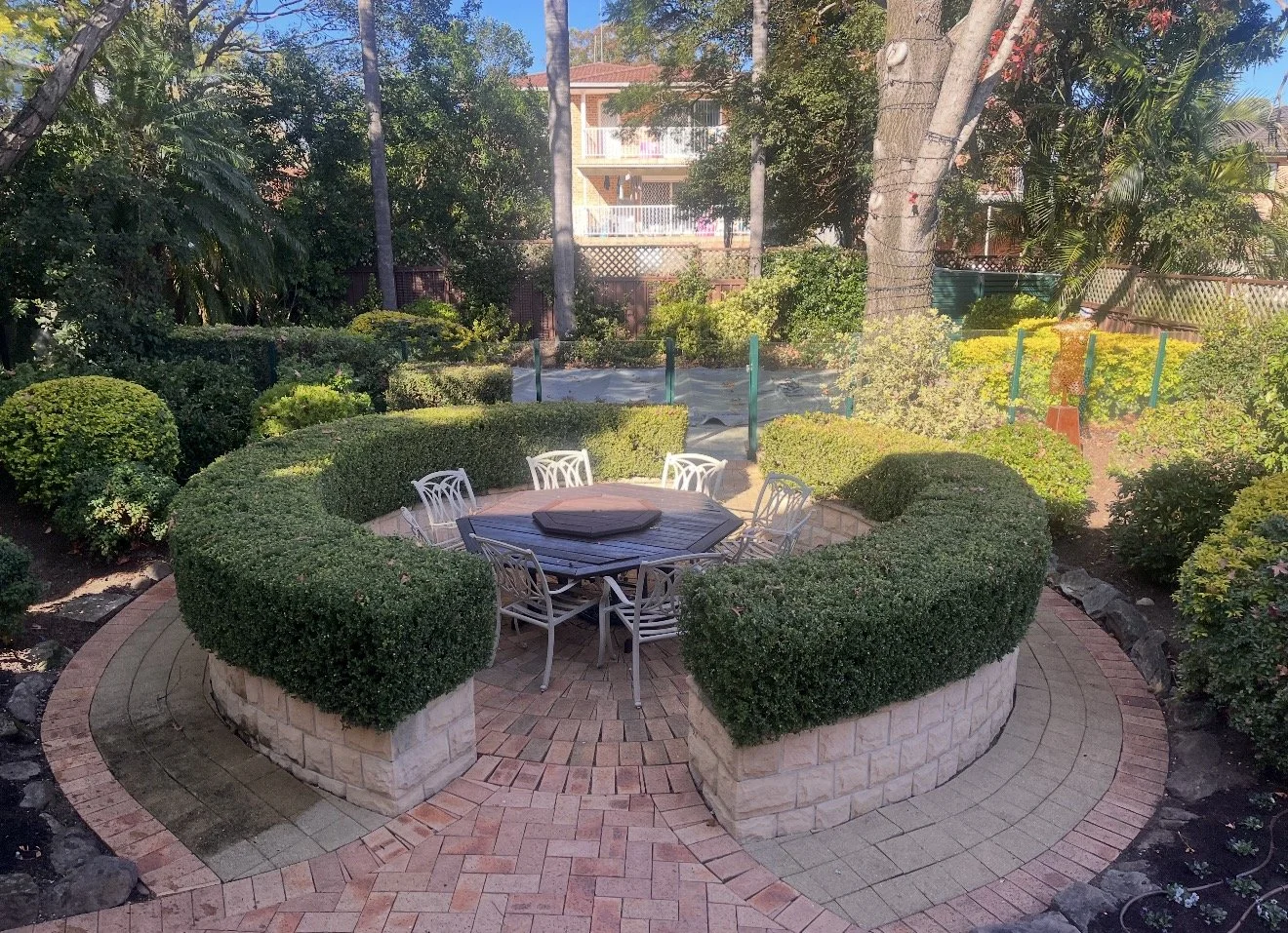 A circular brick patio with a table and six white chairs surrounded by neatly trimmed bushes in a garden.