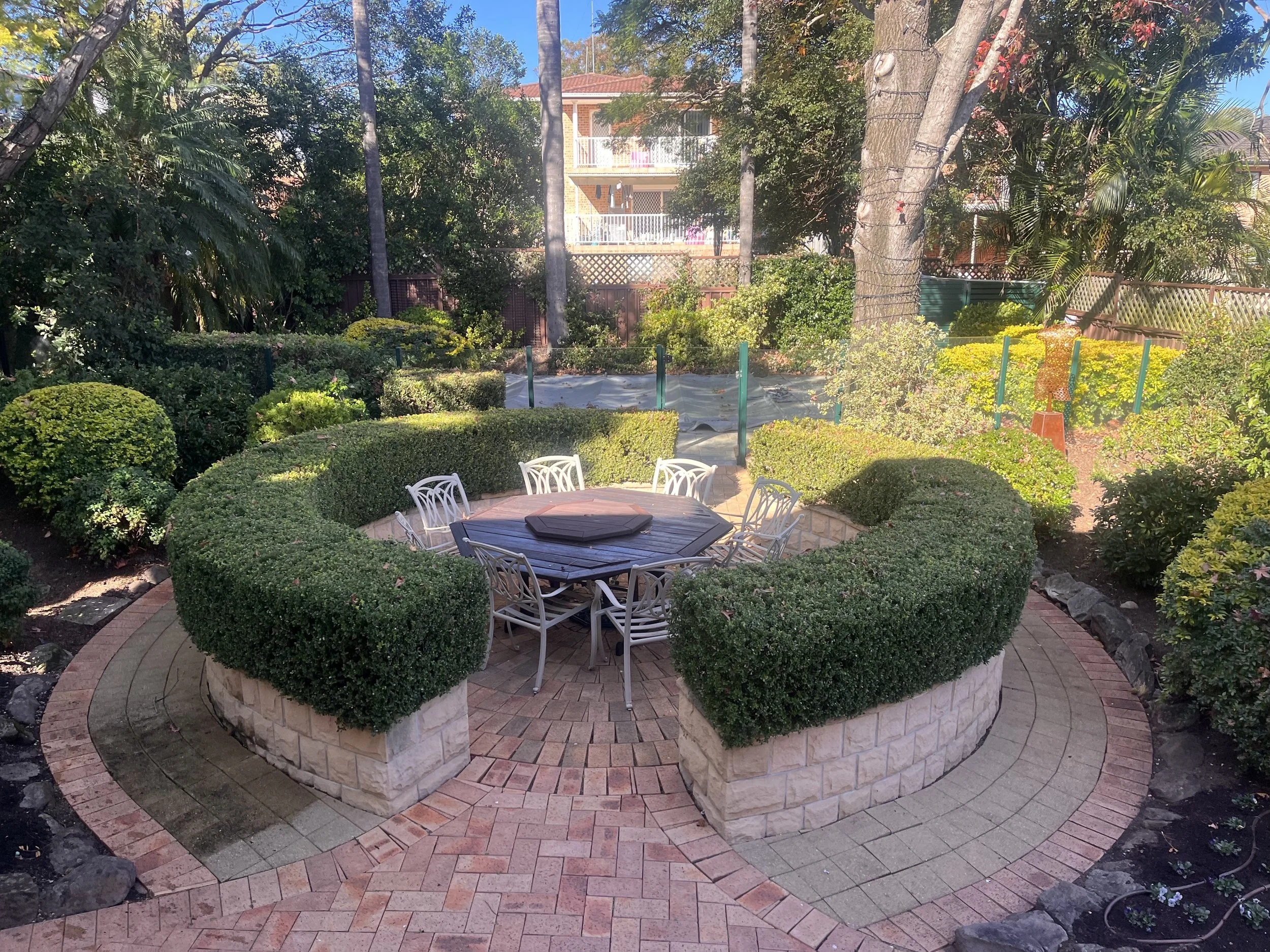 A backyard garden with a round brick patio and white outdoor chairs surrounding a round table with a dark cover. The garden is lush with green bushes, trees, and plants, and there is a small walkway made of bricks running around the circular garden b