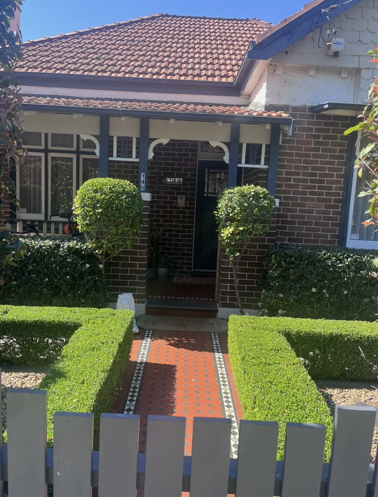 Front view of a brick house with a tiled roof, a small garden with trimmed hedges, two small trees, and a pathway leading to the front door.