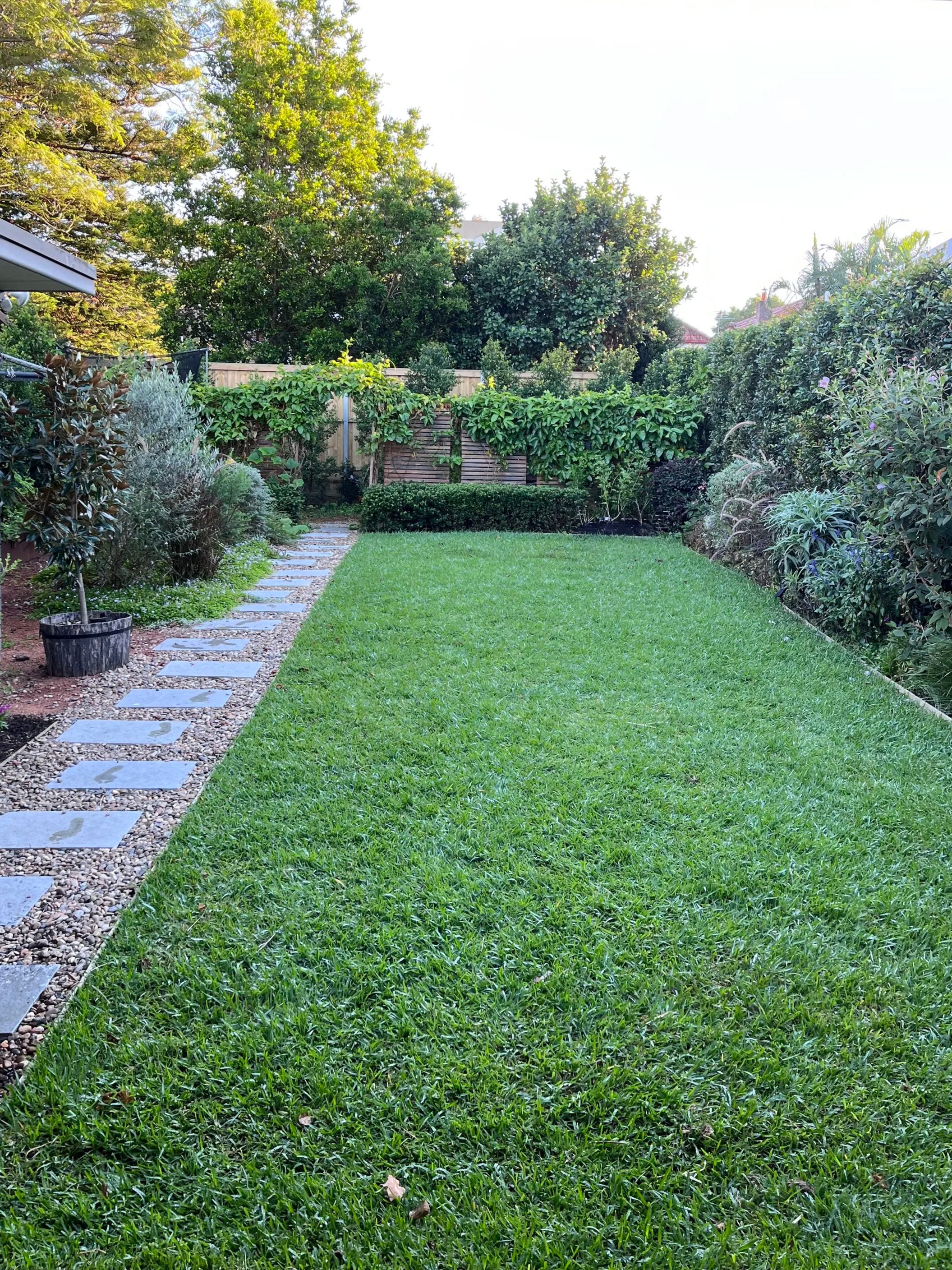 A well-maintained backyard garden with a lush green lawn, a stone pathway on the left, various shrubs and plants, and tall trees in the background. A wooden fence is partially visible behind the greenery.