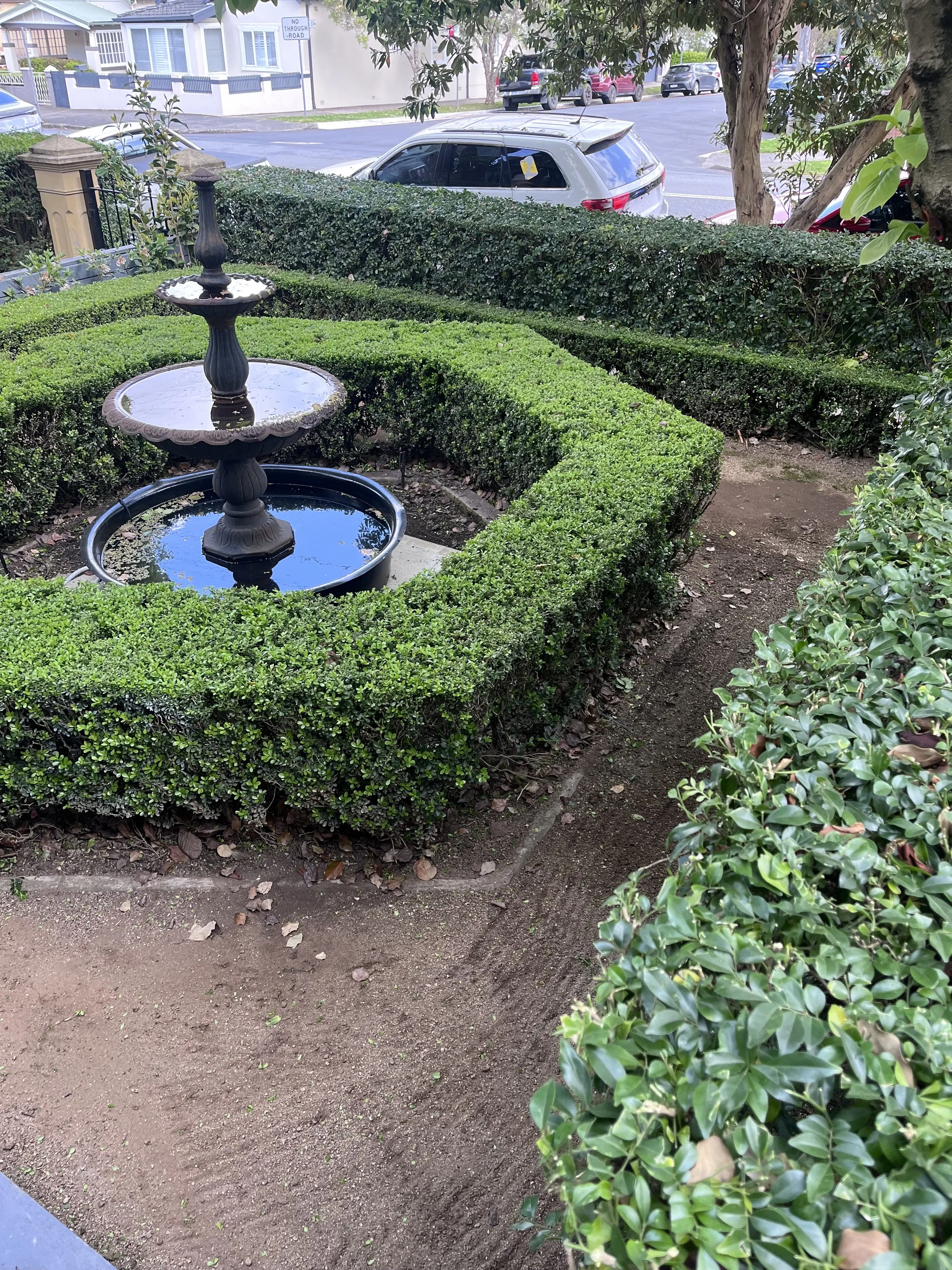 A small garden with a tiered fountain and trimmed bushes, located near a sidewalk and street with parked cars.