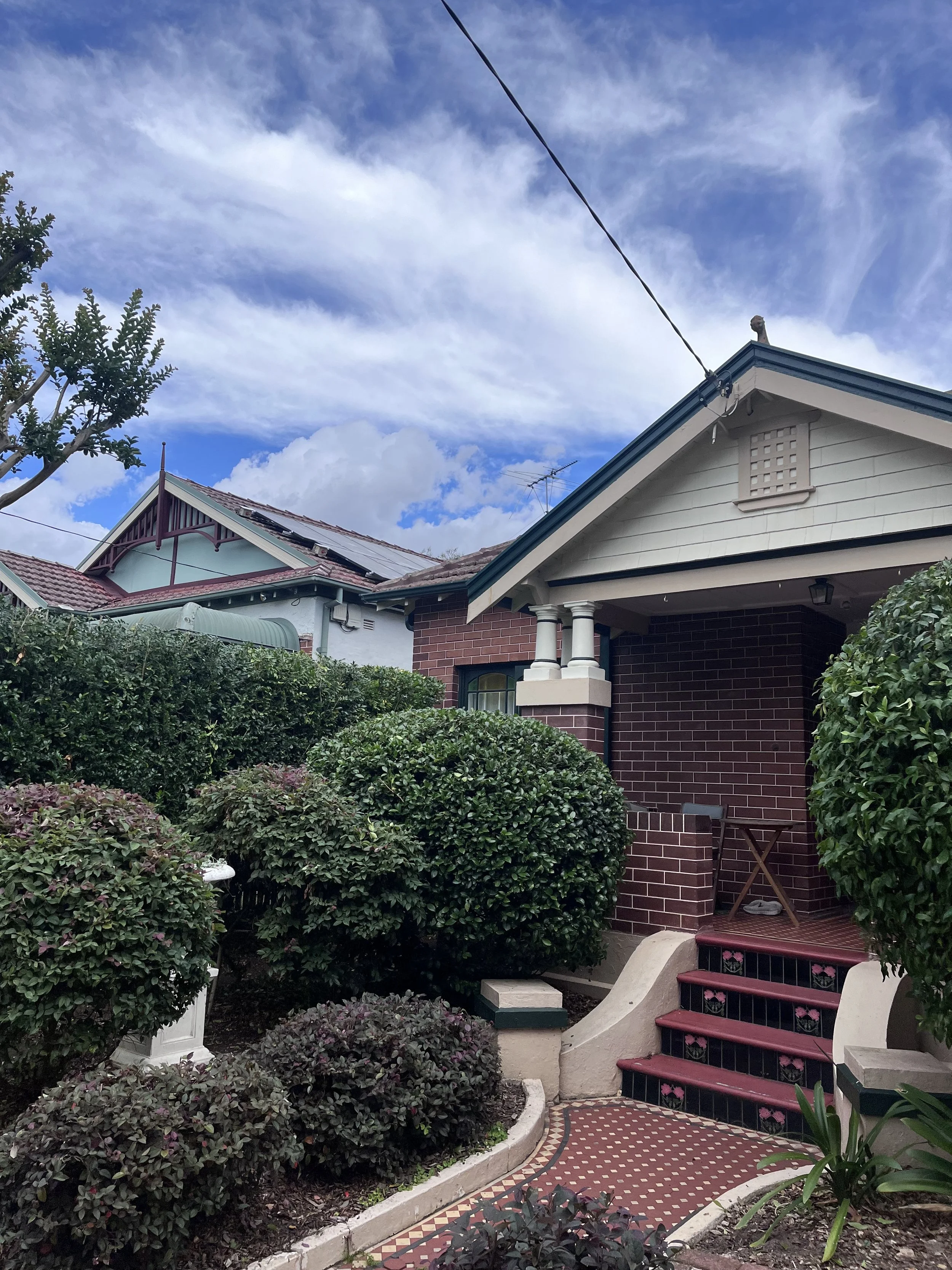 Front porch of a house with stairs, surrounded by lush bushes and plants, under a partly cloudy sky.