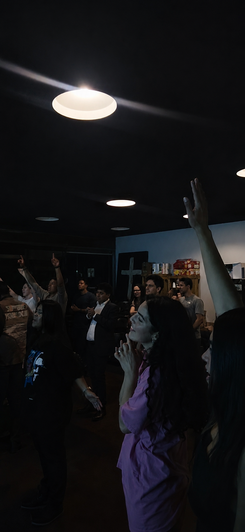 A group of people gathered in a dark room, some raising their hands and others standing with attentive expressions, with ceiling lights above and shelves with boxes and a cross in the background.