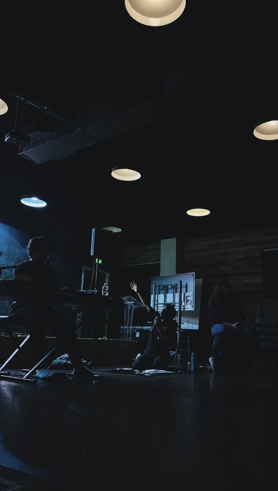 A dark indoor space with three people, one sitting at a keyboard, another kneeling on the floor with a raised hand, and a third seated to the right. There are ceiling lights and a basketball hoop in the background.