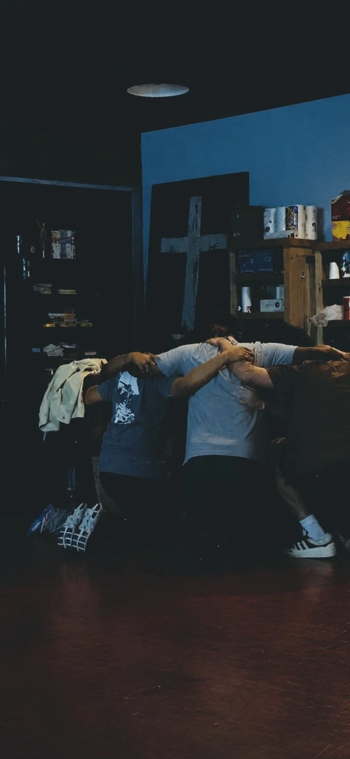 Group of people kneeling in front of a cross on the wall, embracing each other in a prayer or moment of reflection in a dimly lit room.